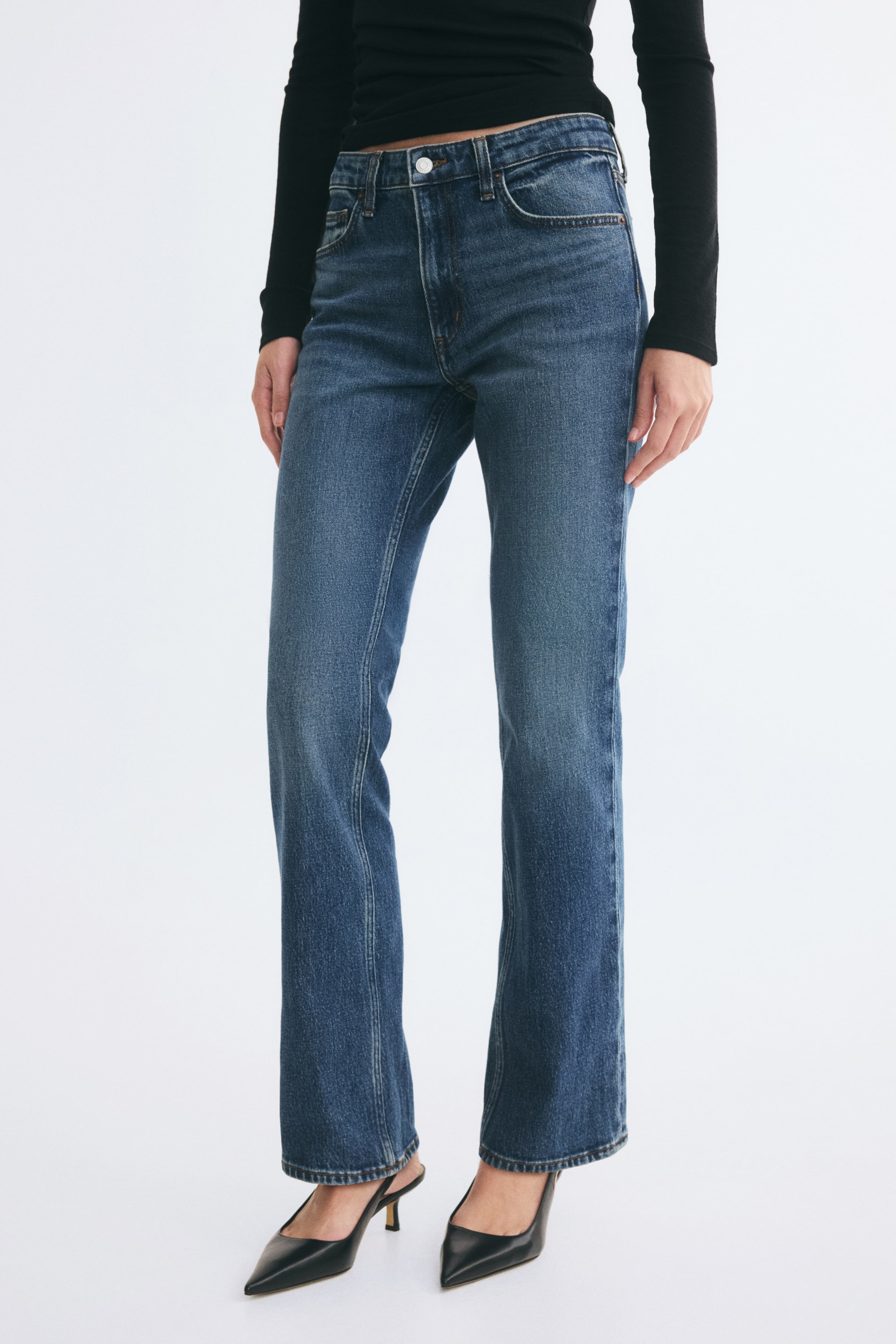 Ver imagen más grande: La modelo lleva unos vaqueros bootcut de denim azul, con cintura regular, corte ajustado en los muslos y la pernera ligeramente acampanada. Los vaqueros se combinan con un top negro de manga larga y zapatos de tacón negros de punta.