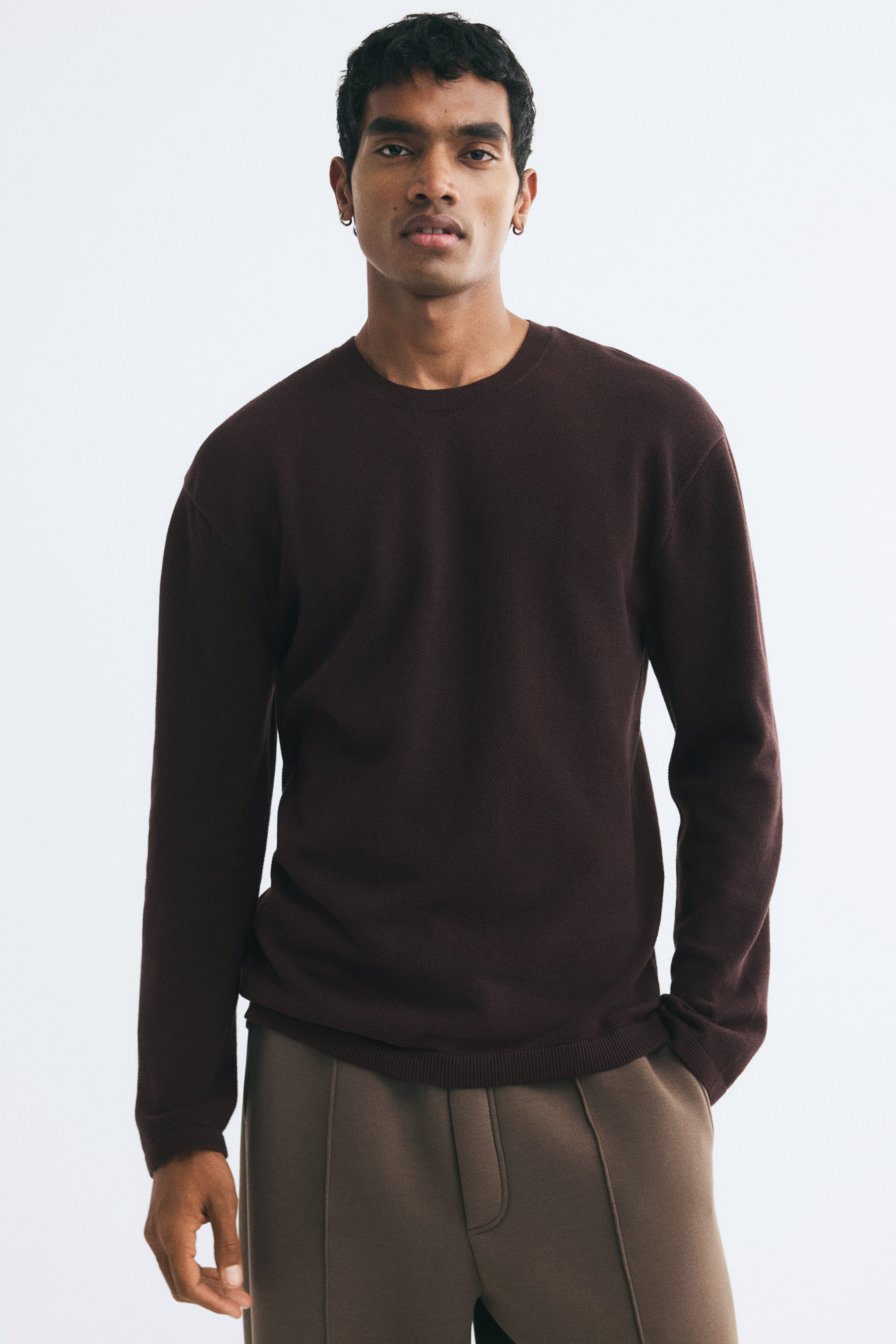 View larger image: A dark brown textured knit sweater is being worn by the model, featuring a relaxed fit, a round neckline, and ribbing at the collar, cuffs, and hem. The sweater is paired with light brown trousers, with the model resting one hand in a pocket.