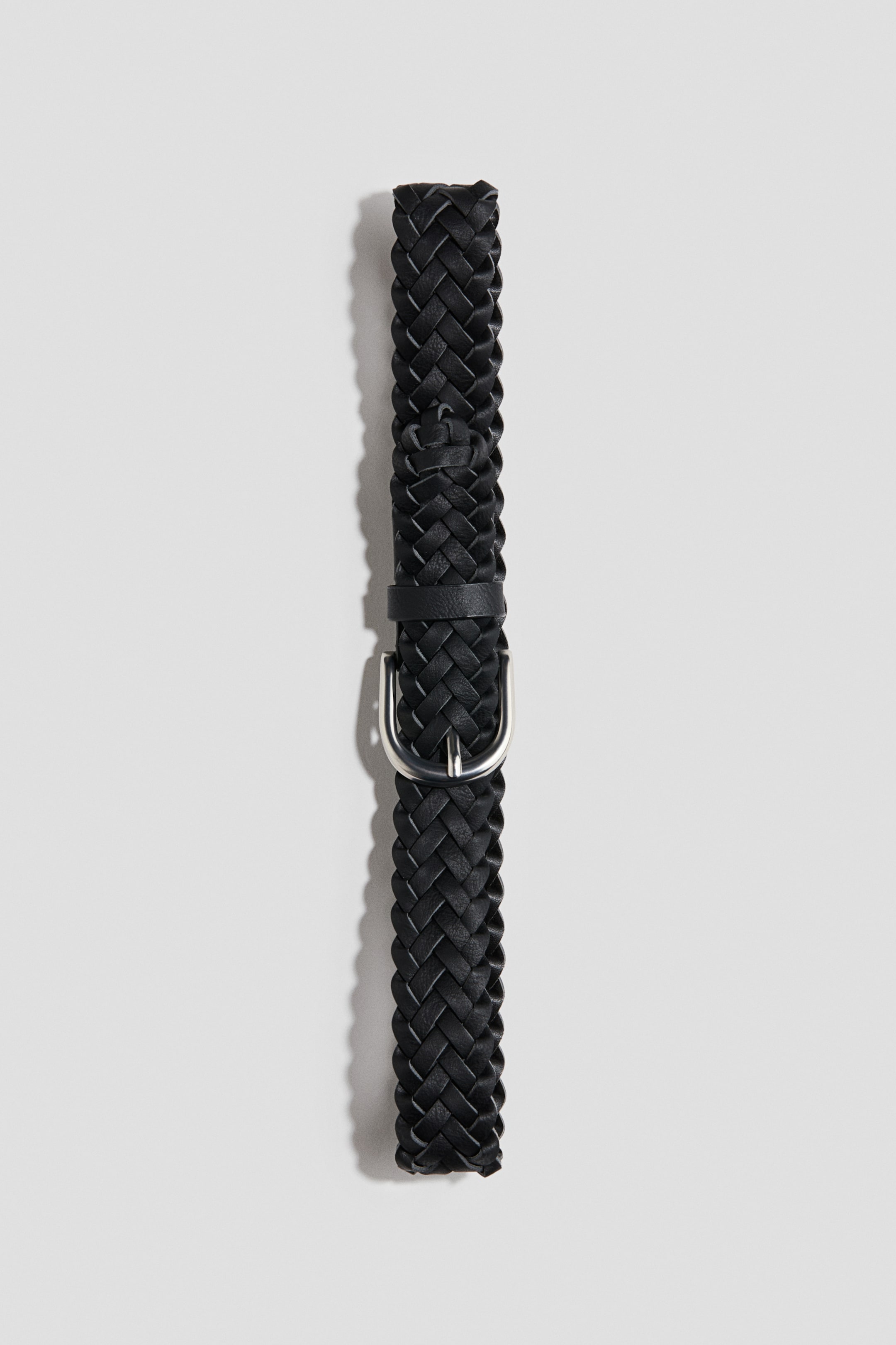 Agrandir l'image: Une ceinture tressée noire est posée verticalement, orientée de face pour mettre en valeur son design. Elle présente une sangle finement tissée, un passant en cuir assorti et une boucle métallique arrondie de ton argenté.