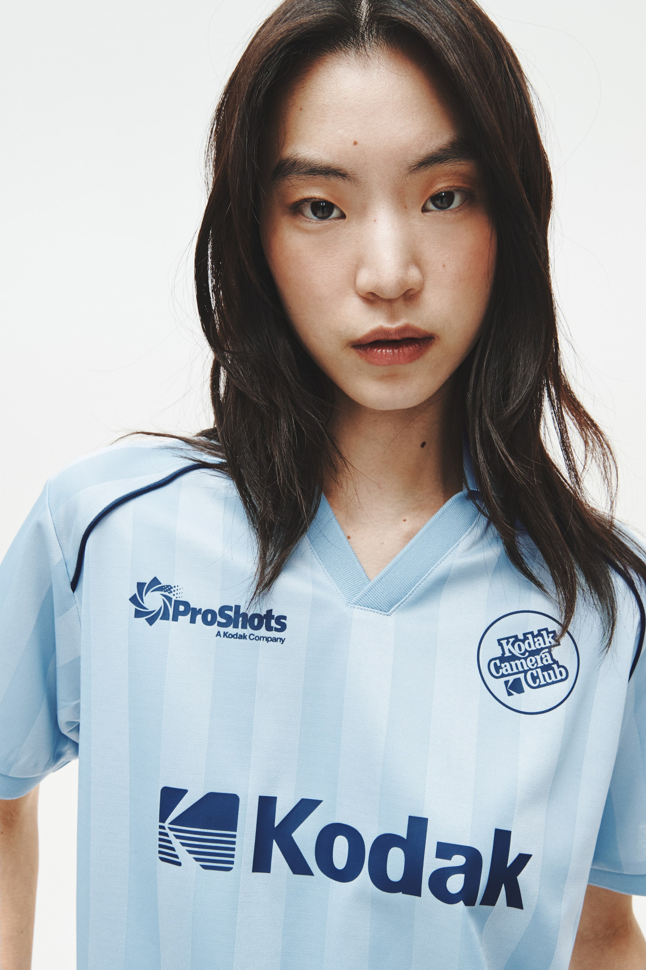 View larger image: A light blue short-sleeved jersey featuring subtle vertical stripes and a ribbed V-neck collar. Dark blue logos for 'ProShots', 'Kodak Camera Club', and 'Kodak' are prominently displayed on the front, with dark blue piping detailing the shoulder seam.