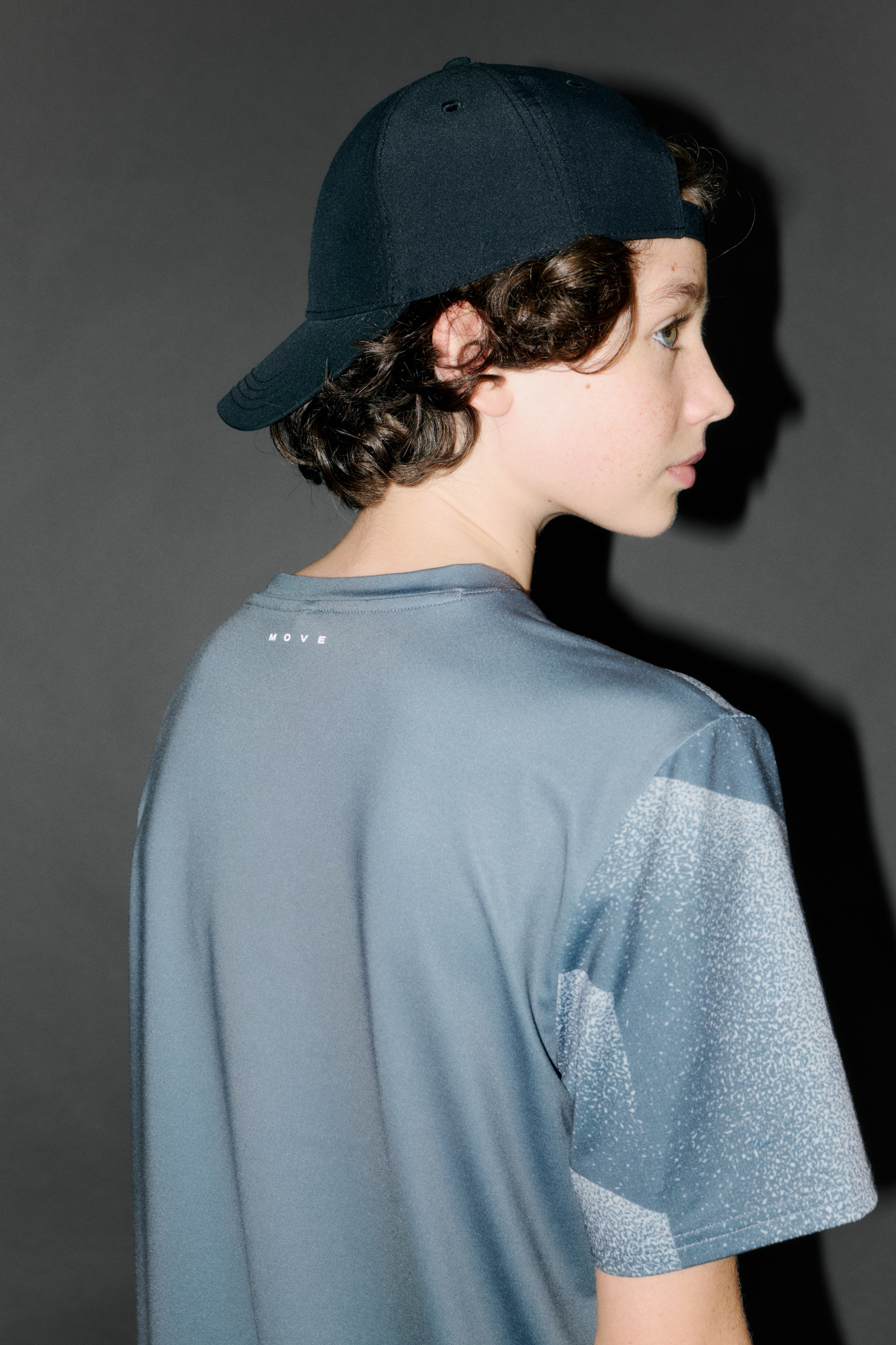 View larger image: A blue-gray short-sleeved t-shirt is being presented from a back and side angle, featuring a light speckled pattern forming geometric shapes on the sleeves and upper shoulder areas, with the word "MOVE" printed in white on the back neckline. A dark baseball cap is also being shown, covering the back of the head.
