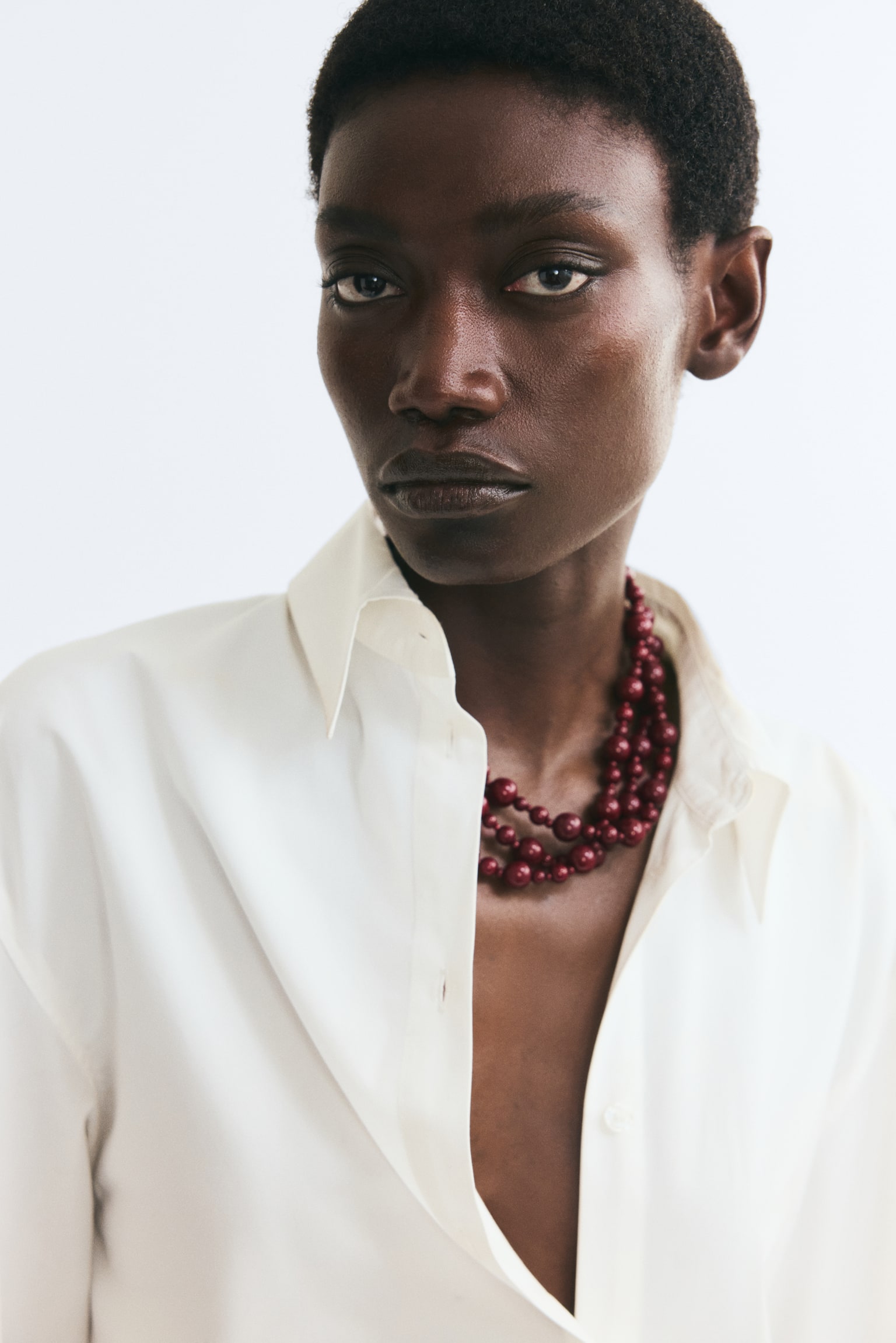 A triple-strand necklace featuring glossy, dark red beads of varying sizes is being worn by the model. The layered necklace is styled under an open-collared white shirt, showcasing how the strands drape against the chest. - 1
