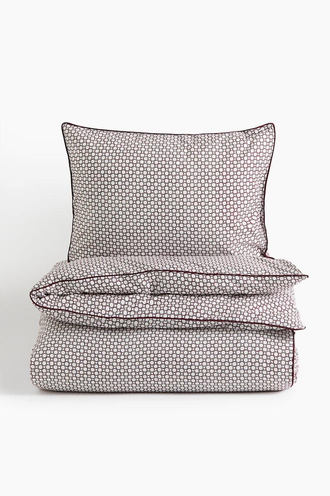 A bedding set featuring a matching duvet cover and pillowcase with a burgundy and white geometric grid pattern is shown from the front. The folded duvet cover is stacked neatly under the upright pillowcase, with both items displaying thin burgundy piping along their edges. - 1