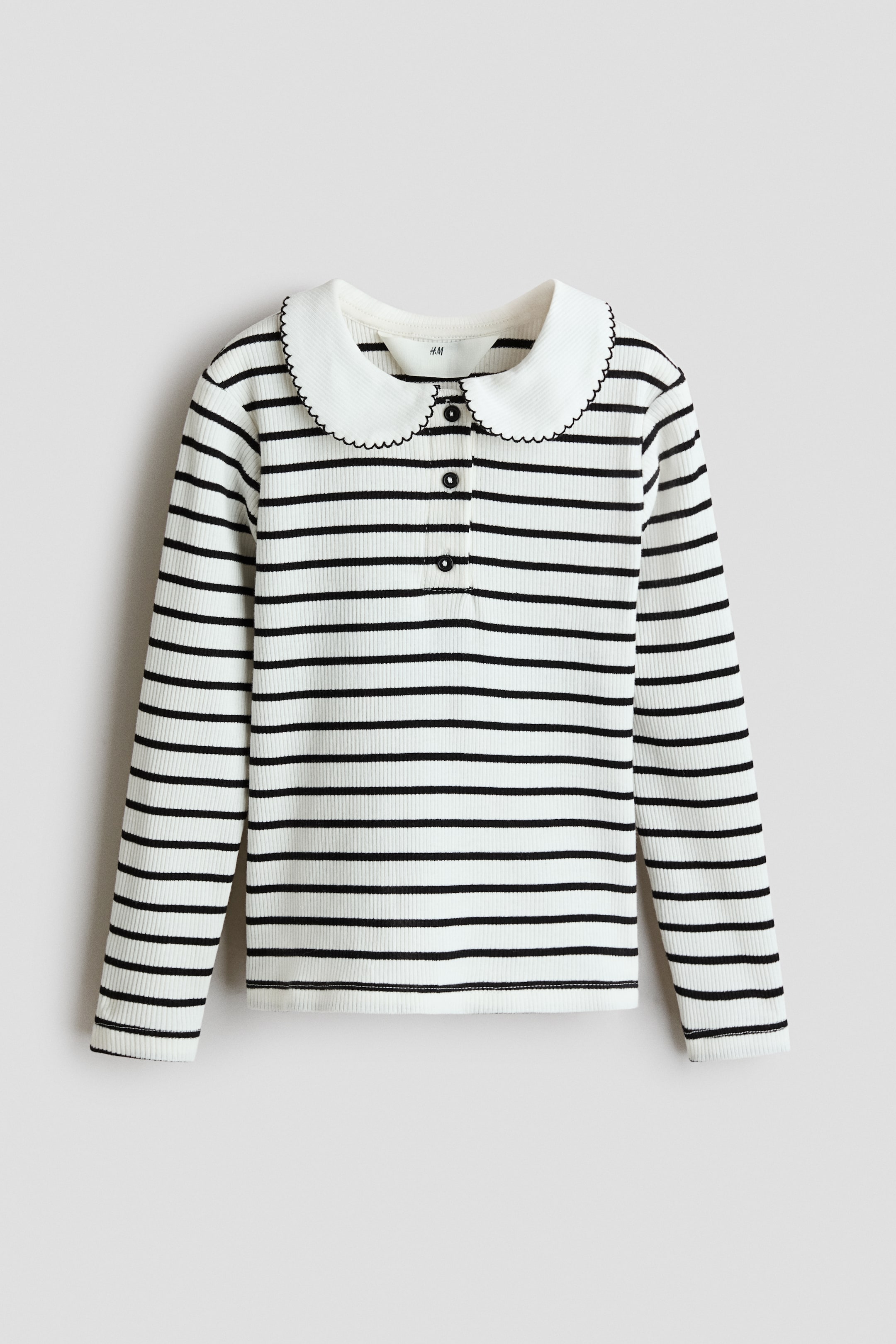 View larger image: A long-sleeved, front-facing ribbed top features a white and black horizontal stripe pattern. It has a white rounded Peter Pan collar with a black scalloped edge and a three-button placket at the chest.