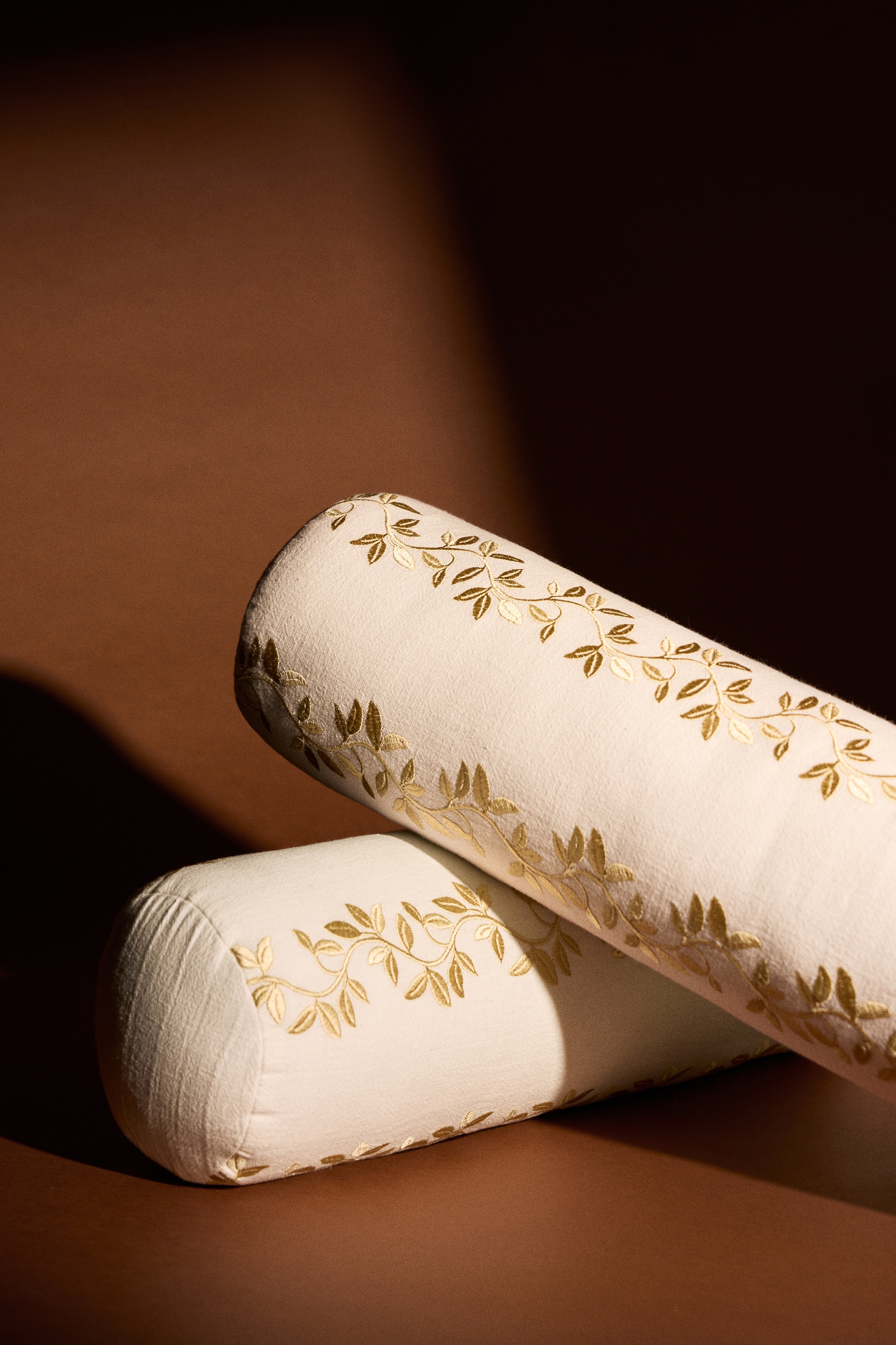 A cylindrical bolster pillow crafted from off-white cotton and linen canvas is lying horizontally, showcasing a gold satin-stitched leafy vine motif running across its front. The bolster features rounded ends and a visible woven texture, with the detailed embroidery accentuating its elongated shape.