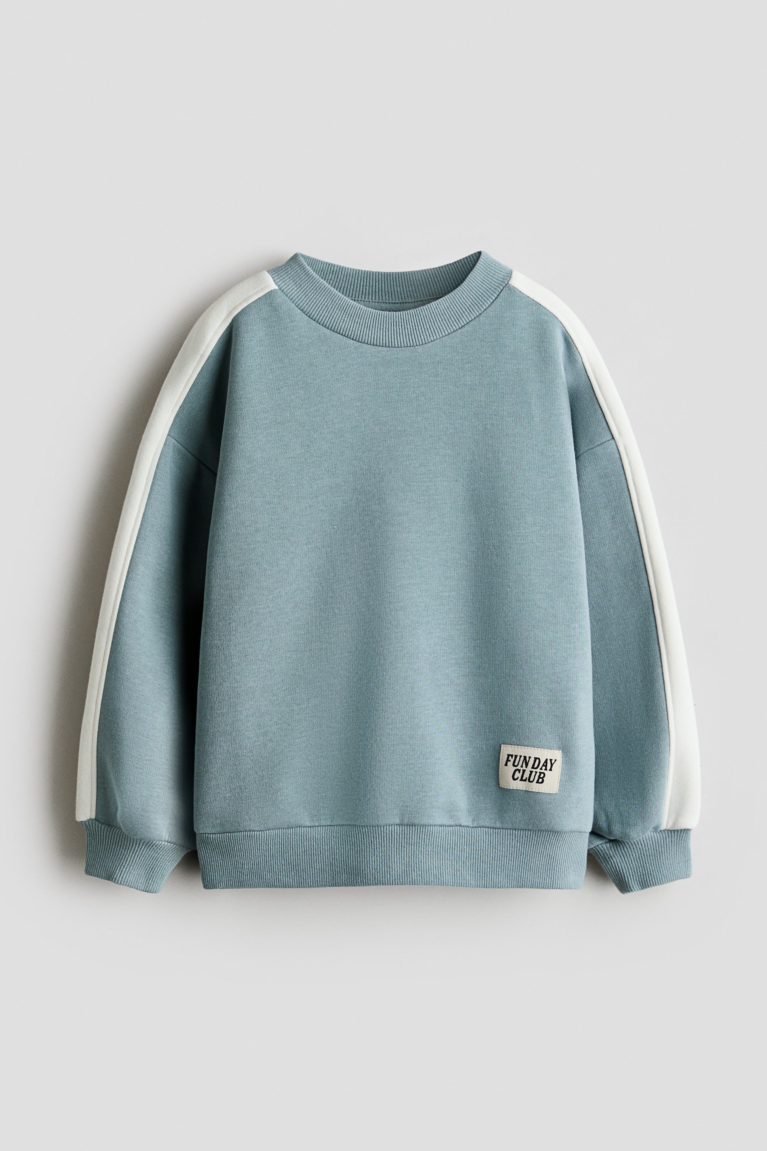 Un sweat-shirt coupe ample en tissu polaire bleu-gris attÃ©nuÃ© est prÃ©sentÃ© de face, avec un col rond bordÃ© de bord-cÃ´te, des Ã©paules tombantes et des bandes contrastantes blanches courant le long des manches. Le vÃªtement prÃ©sente des poignets et un ourlet cÃ´telÃ©s, avec une petite patch rectangulaire portant lâinscription Â« FUN DAY CLUB Â» dÃ©taillÃ©e sur la partie infÃ©rieure avant gauche.