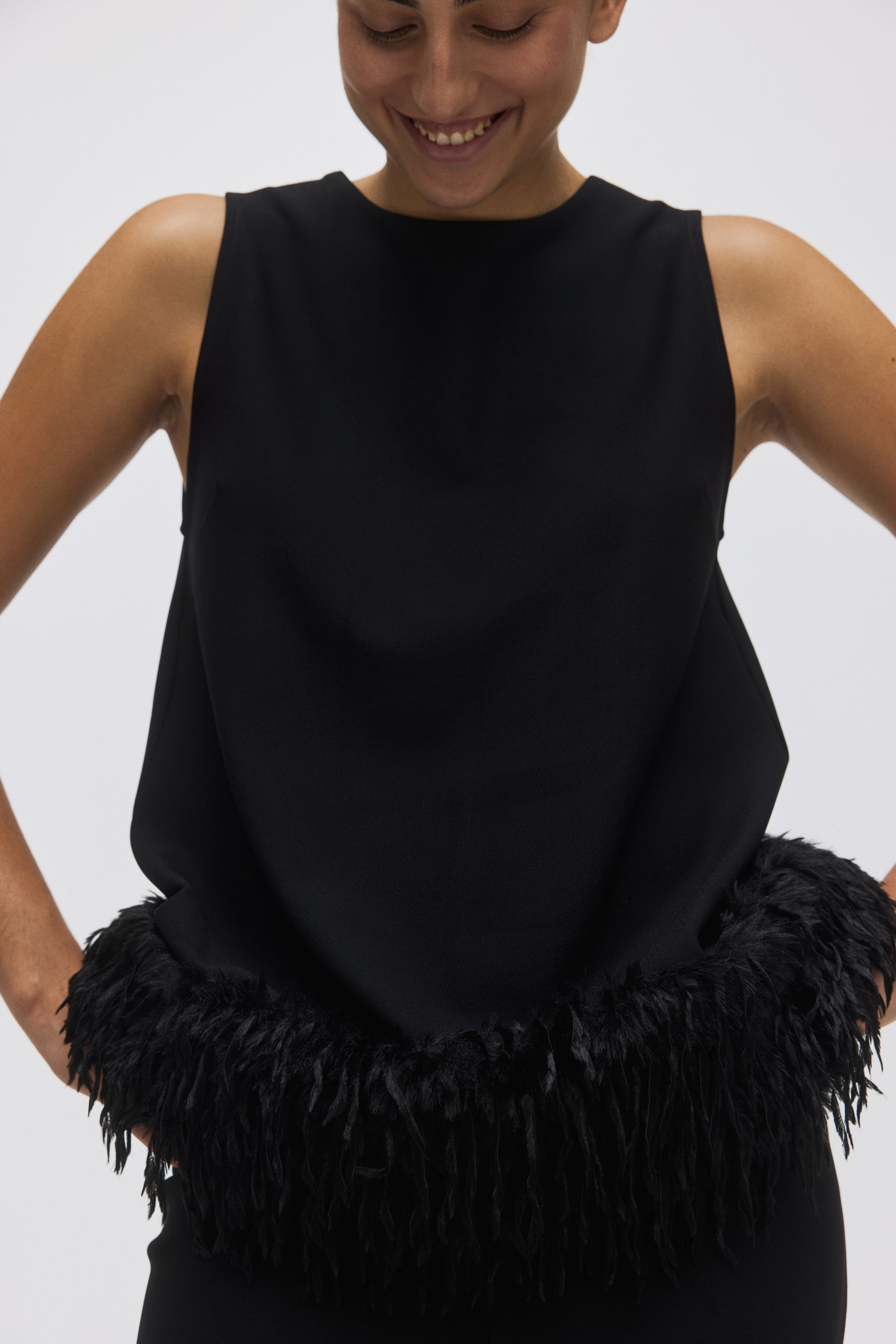 View larger image: A black sleeveless top is being shown from the front. It is featuring a high, round neckline and a relaxed fit, with its bottom hem being embellished with a thick band of black feathers.