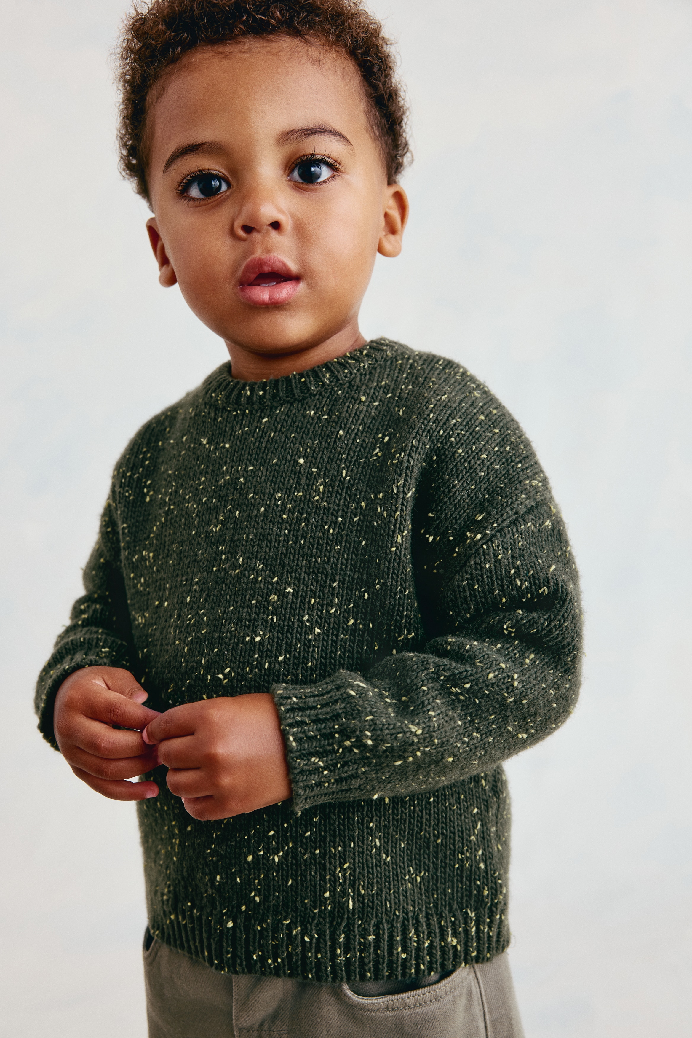The dark green sweater is shown from the front, featuring a textured knit with yellow flecks throughout. It is designed with a ribbed crew neck, ribbed cuffs, and a ribbed hem.