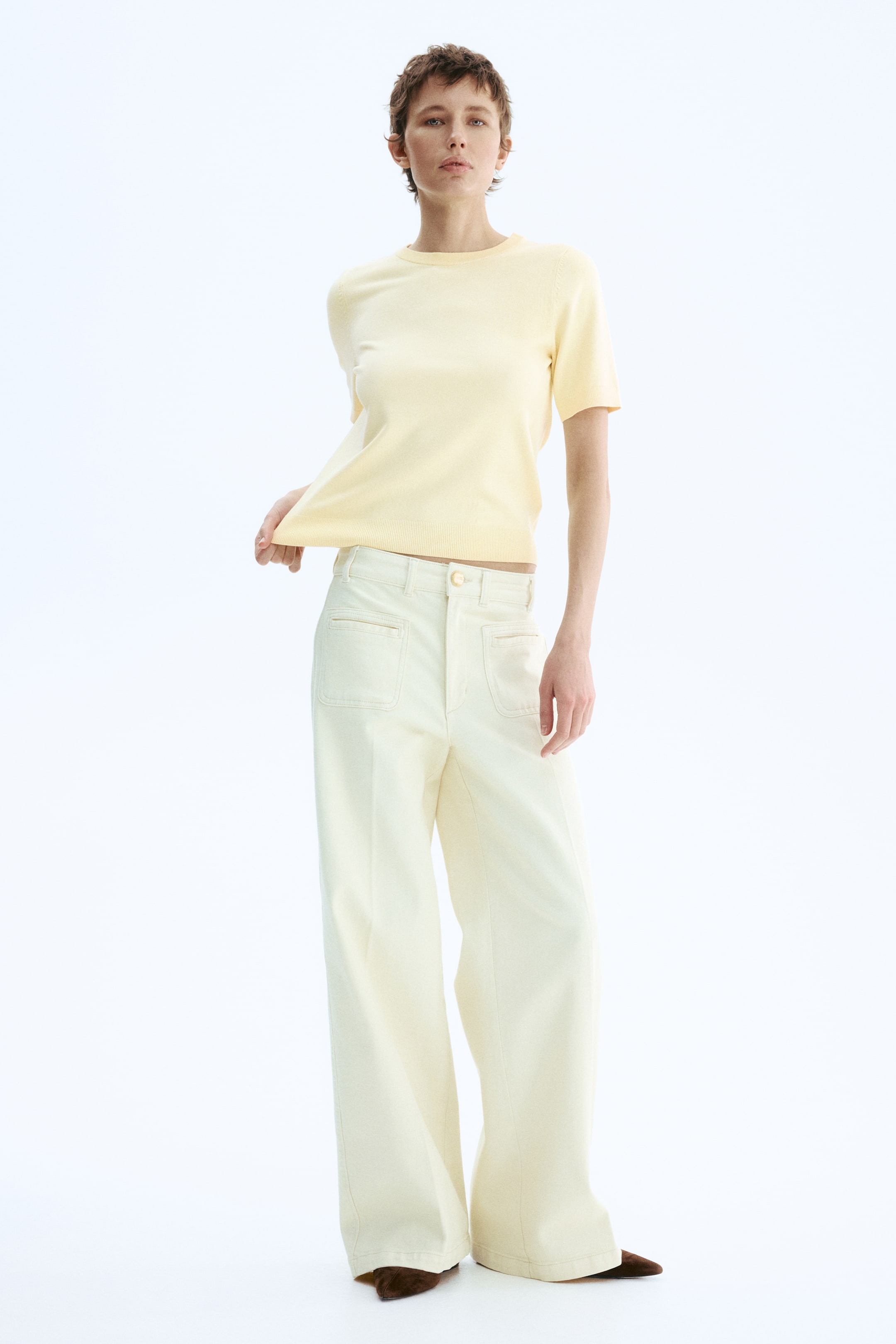 View larger image: The light yellow short-sleeved knit top is showcased from a slightly angled front perspective, highlighting its crew neck and finished bottom hem. It is paired with light cream wide-leg pants, which include front patch pockets, belt loops, and a visible button closure.