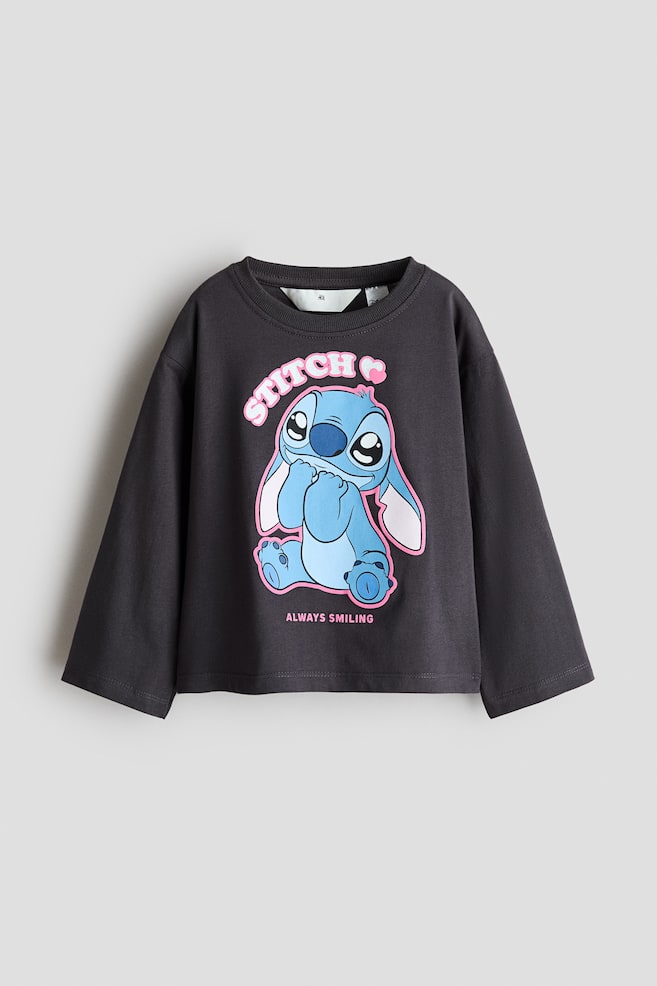 Un t-shirt à manches longues gris foncé est présenté de face, avec un col rond doté dâune fine bordure côtelée et des manches à coupe décontractée. Le devant du t-shirt met en valeur un motif imprimé du personnage de dessin animé Stitch, avec des lettres roses indiquant « STITCH » au-dessus et « ALWAYS SMILING » en dessous. - 1