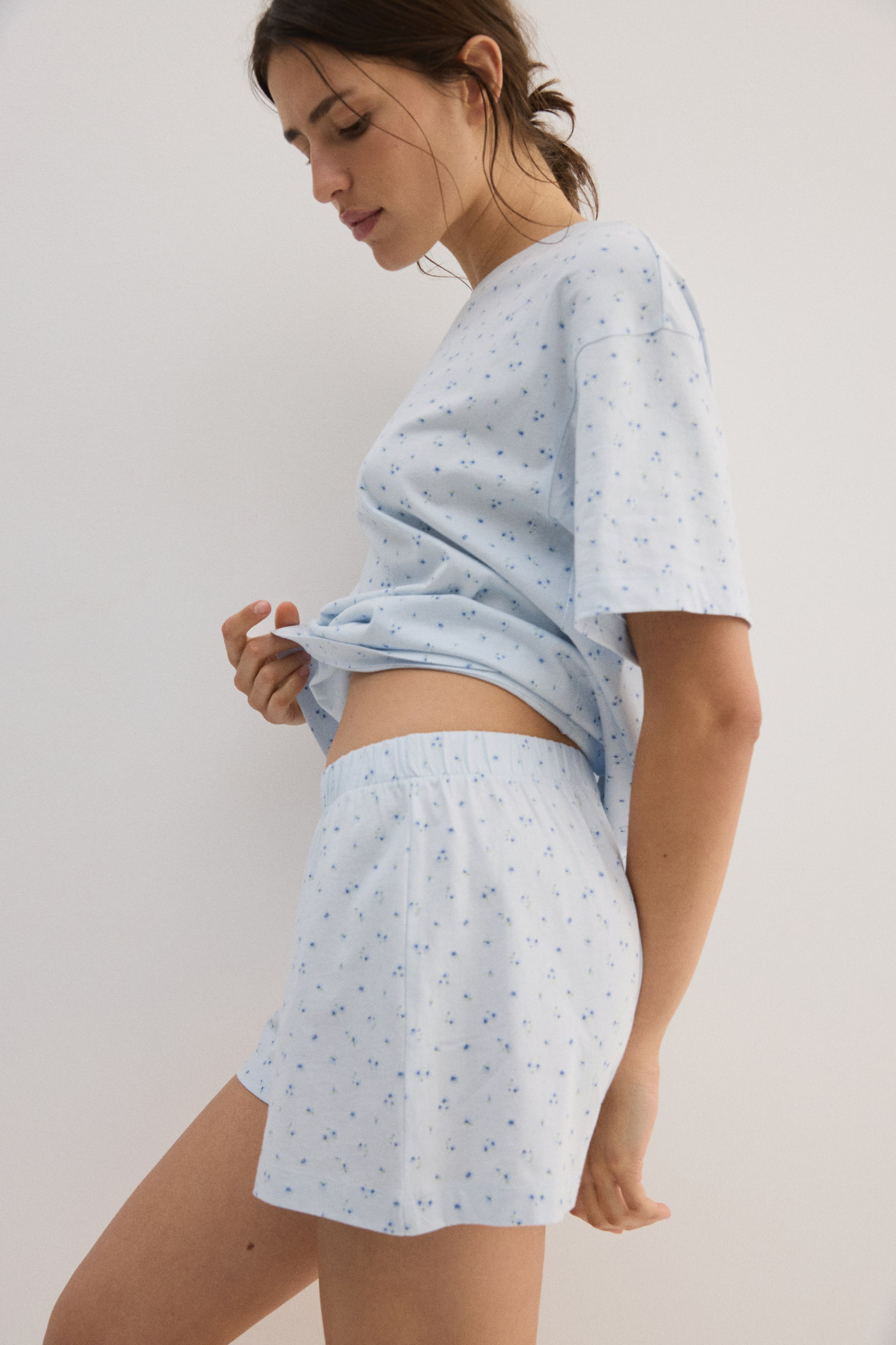 Grotere afbeelding bekijken: Een lichtblauw katoenen jersey pyjamaset, bestaande uit een T-shirt met verlaagde schouders en een geribde halslijn, en shorts met een bedekte elastische tailleband, wordt gedragen door het model. De pyjama's hebben een bedrukt bloemenpatroon en het model tilt de zoom van het T-shirt op, waardoor de tailleband van de shorts zichtbaar wordt.