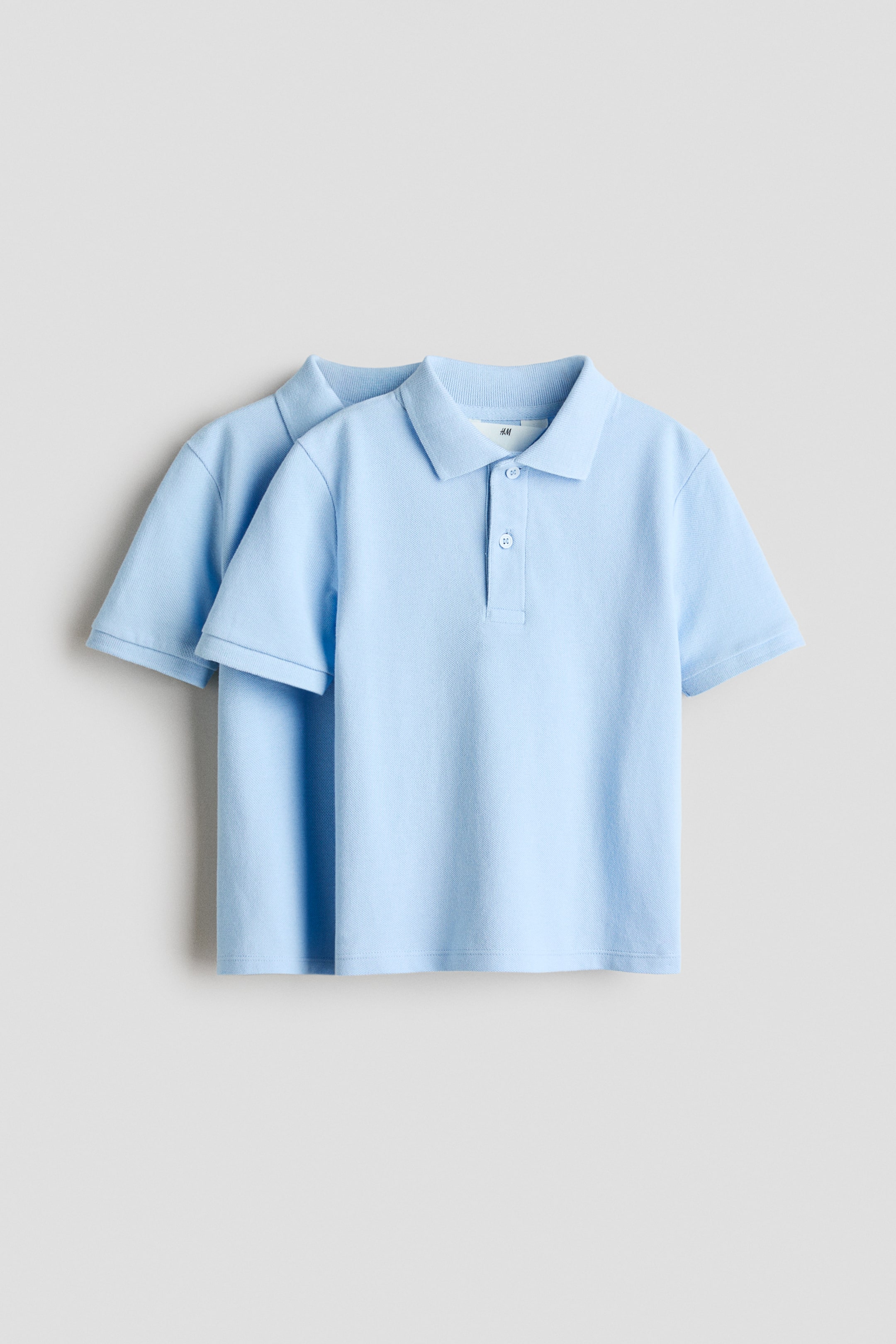 View larger image: Two light blue short-sleeved polo shirts are layered, showcasing their front views with ribbed polo collars and two-button plackets.