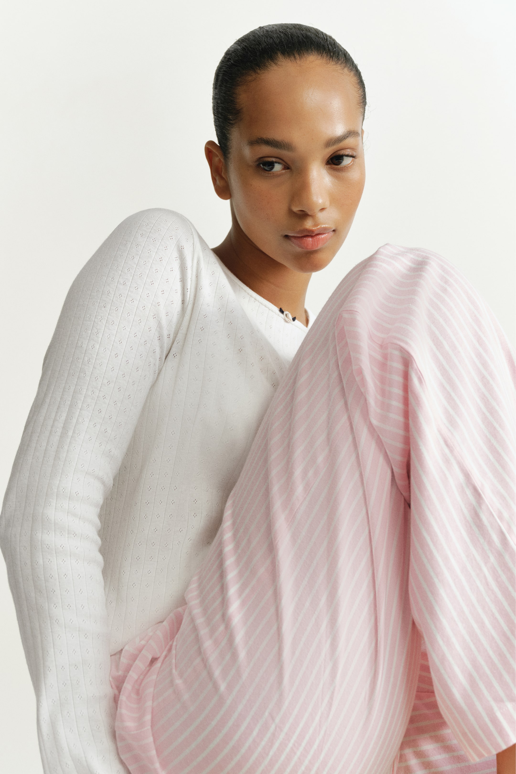 Grotere afbeelding bekijken: De pyjama bestaat uit een witte, lange mouwen top van katoenen pointelle-jersey en een roze-wit gestreepte flanellen broek, gedragen door het model. Het model zit met één knie opgetrokken en laat zo de gestructureerde breistructuur van de top en de zachte stof van de gestreepte broek zien.
