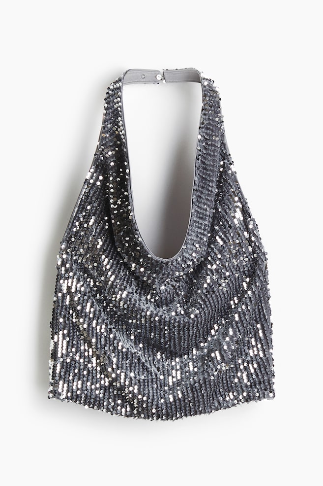 A silver sequined halter-neck top is hanging, presenting its front. The garment features a draped neckline and a narrow, grey neck strap secured by two snap buttons, with countless small silver sequins covering its entire shimmering surface. - 1