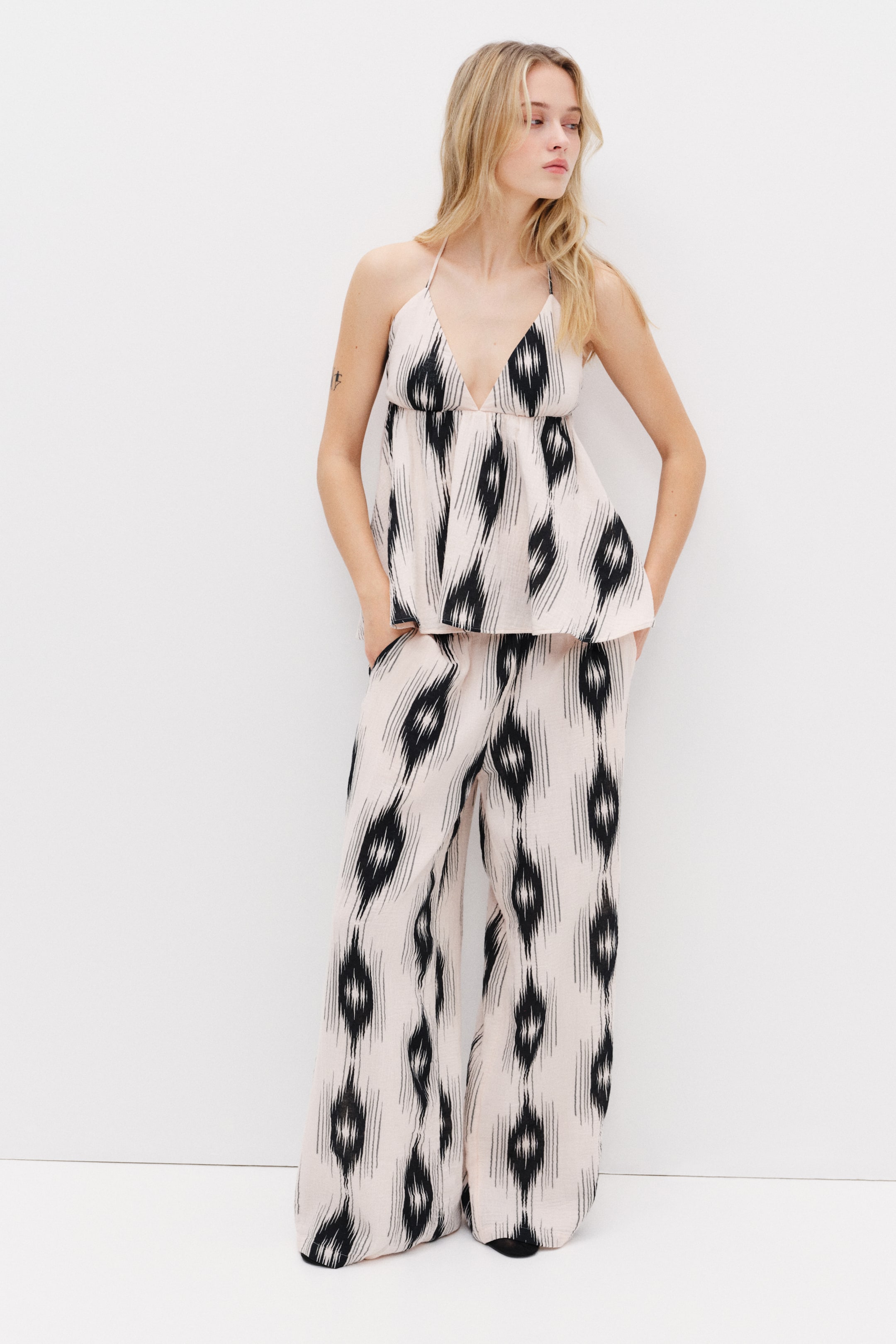 View larger image: Trousers in soft cotton muslin with a light background and black abstract patterns, featuring an elasticated waist, diagonal side pockets, and straight legs, are worn by the model. The model places her hands into the diagonal side pockets, and the trousers are styled with a matching halter neck top.