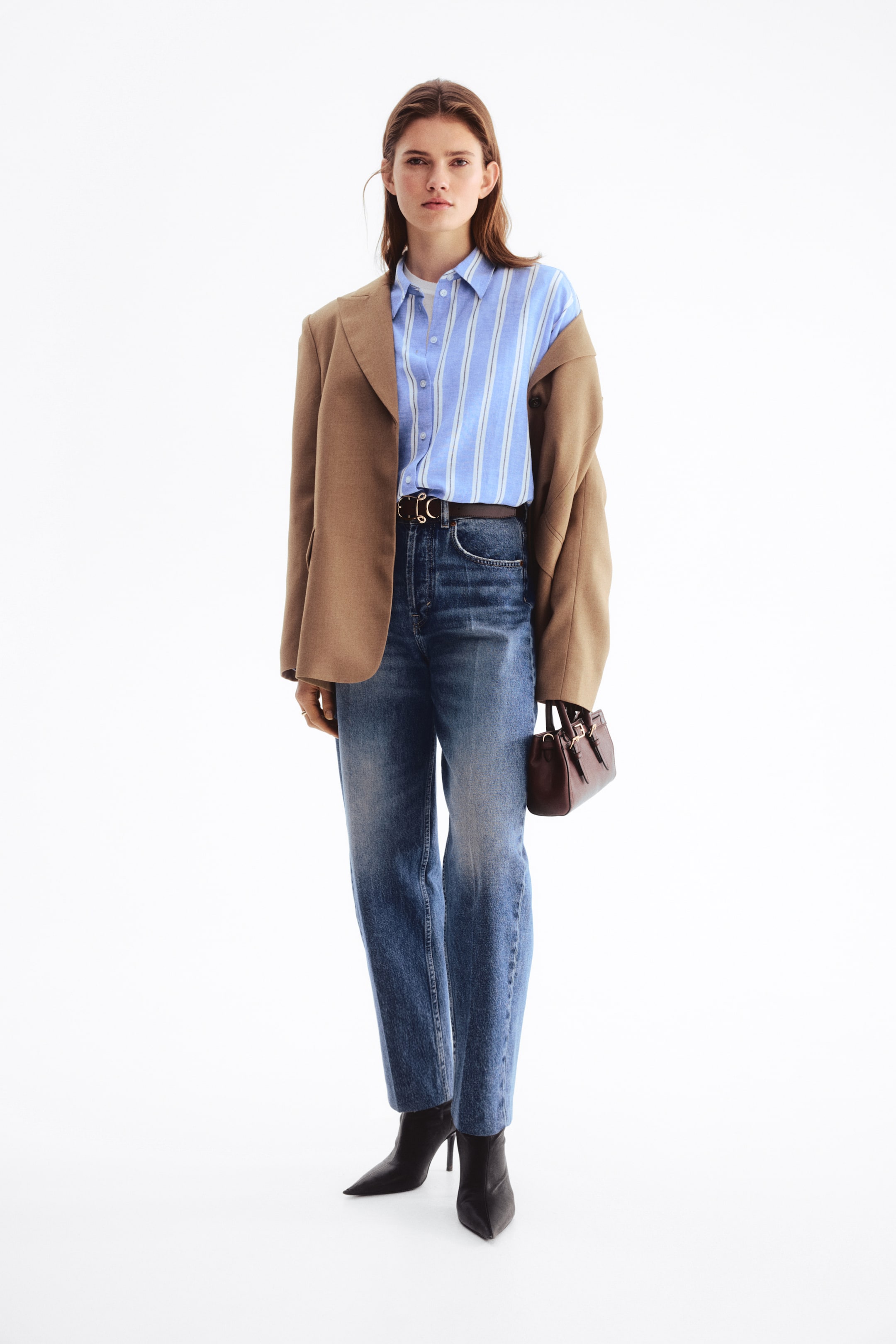 Vis større bilde: En lys blå stripete skjorte med knapper er stukket inn i høy midje blå jeans med rette ben og rå kant, med en brun blazer drapert over skuldrene. Et mørkebrunt belte med et gullfarget dobbeltring-spenne strammer inn midjen, mens svarte spisse ankelstøvler fullfører antrekket, og en liten burgunder strukturert håndveske holdes.
