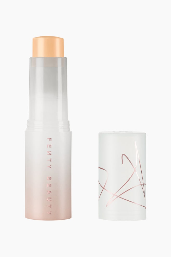 Eaze Drop Blur + Smooth Tint Stick stÃ¥r oprejst og viser en fast, lys ferskenfarvet makeup-formel, der er trukket ud af et matteret, halvtransparent hylster med en pink-til-hvid gradient og lodret "FENTY BEAUTY"-tekst. En matchende cylindrisk hÃ¦tte stÃ¥r til hÃ¸jre og har abstrakte, tynde roseguld-linjer, der er viklet omkring den matterede hvide overflade. - 1