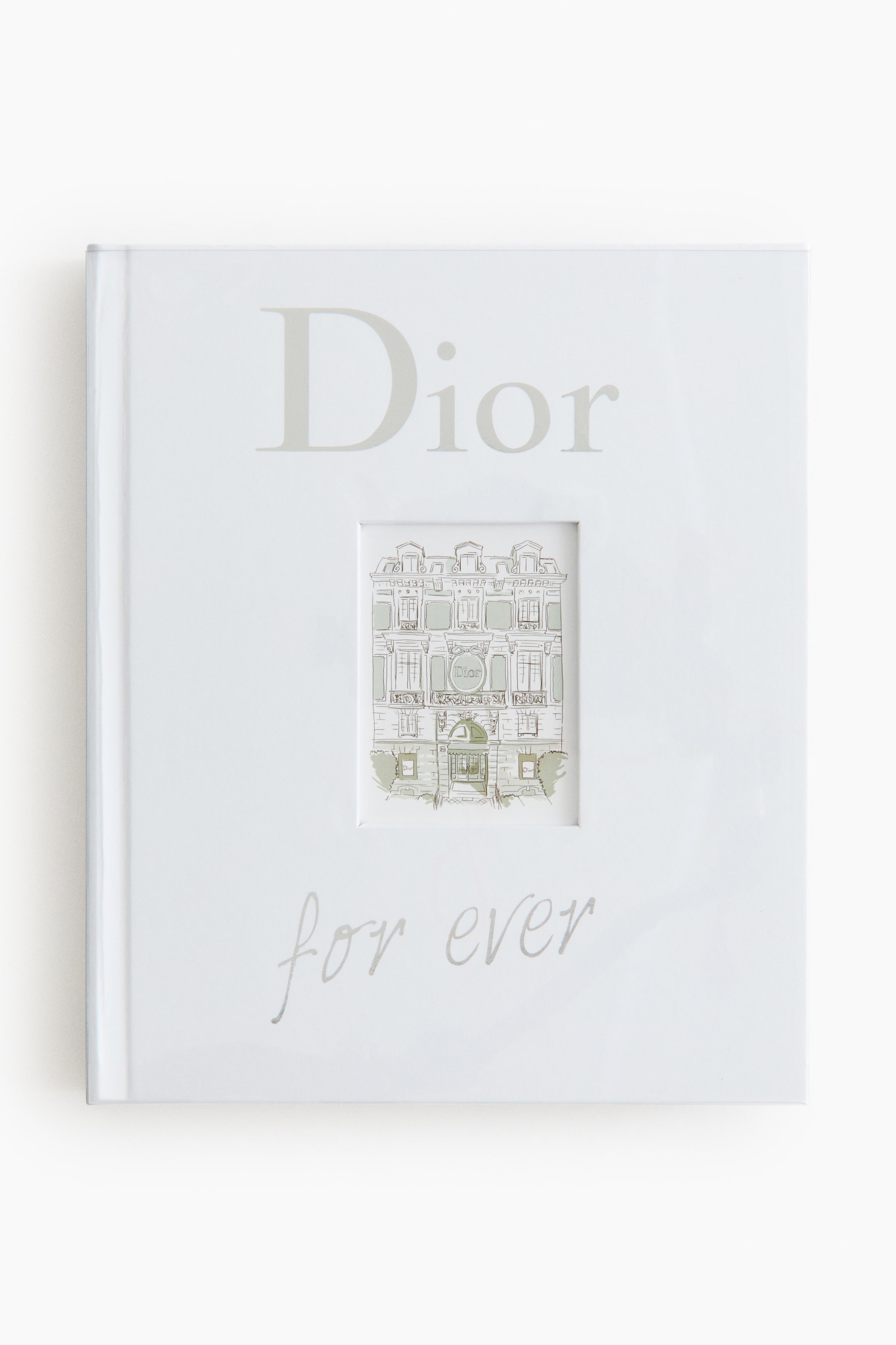 Grotere afbeelding bekijken: Een wit boek wordt VOOR getoond, met "Dior" in lichtgrijze schreefloze tekst bovenaan en "for ever" in lichtgrijze scripttekst onderaan. Een rechthoekige uitsnede in het midden onthult een gedetailleerde grijze en groene architecturale illustratie van een gebouw met meerdere verdiepingen.