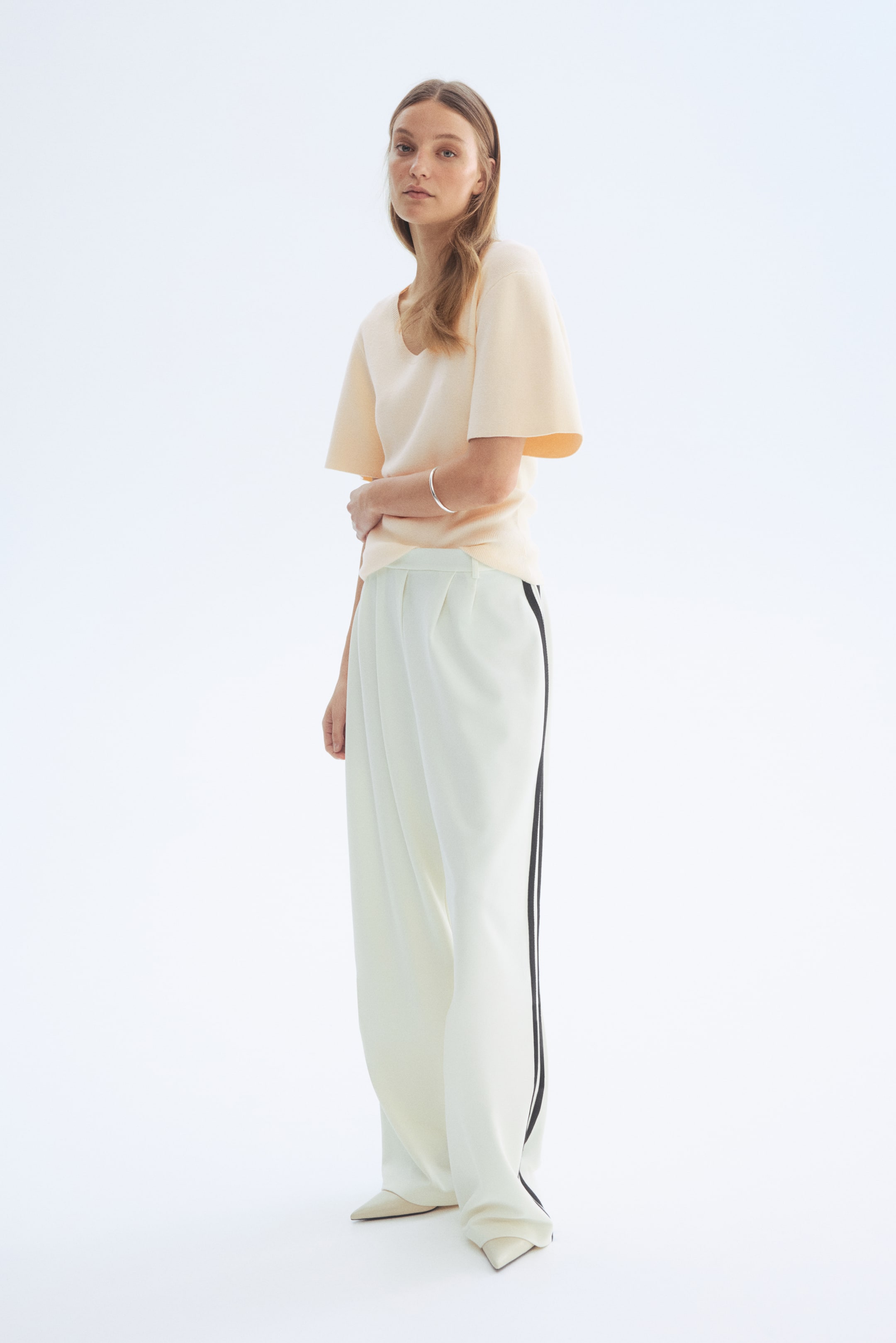 Προβολή μεγαλύτερης εικόνας: Ένα κοντομάνικο τοπ σε ανοιχτό ροδακινί χρώμα με V λαιμόκοψη και φαρδιά μανίκια. Συνδυάζεται με κρεμ παντελόνι με φαρδιά γραμμή, το οποίο διαθέτει πιέτες μπροστά και μια μαύρη ρίγα κατά μήκος κάθε πλευρικής ραφής, ενώ οι μυτερές γόβες είναι ορατές κάτω από το στρίφωμα.