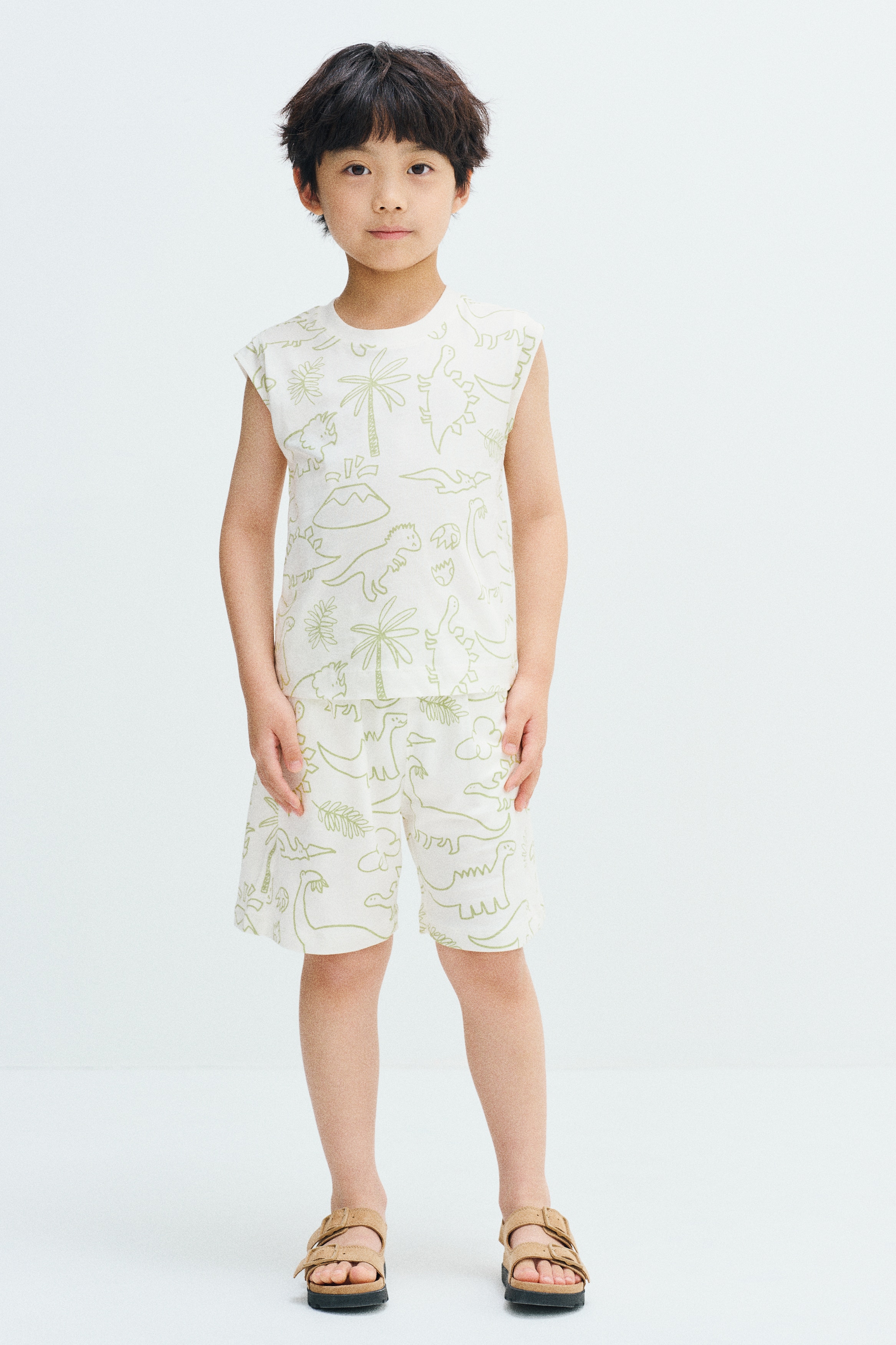 Knee-length shorts in white printed fleece sweatshirt fabric are shown from the front, featuring an elasticated waistband and discreet side pockets. The shorts are decorated with an all-over light green line-art pattern of dinosaurs, clouds, and leaves.