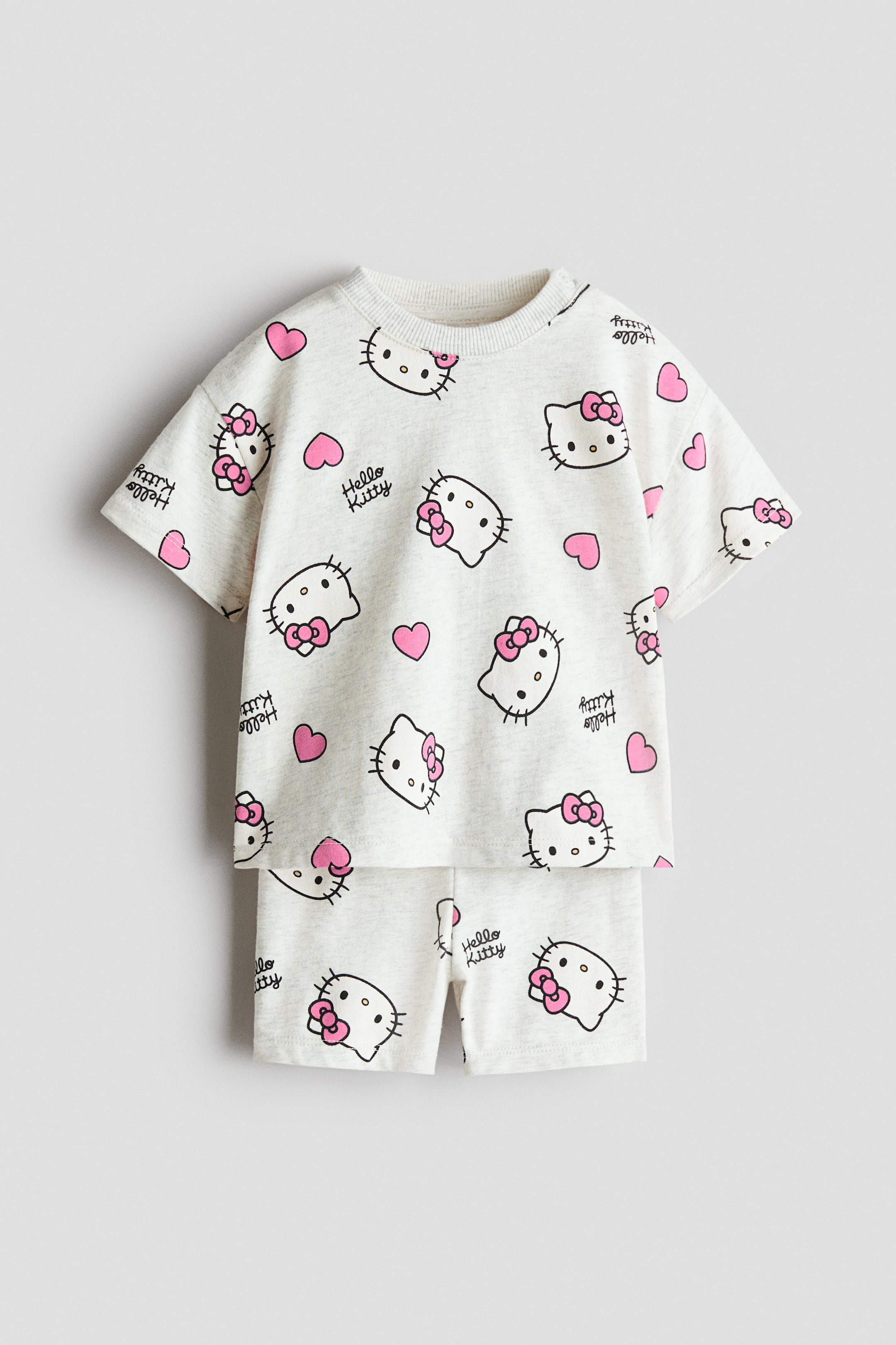Xem ảnh lớn hơn: Bộ đồ cotton in 2 mảnh đang hiển thị mặt trước của nó, bao gồm một chiếc áo thun ngắn tay màu xám nhạt và quần short đồng bộ. Áo có cổ tròn gân và vai rớt, cả hai mảnh đều có họa tiết in toàn bộ khuôn mặt Hello Kitty với nơ hồng, trái tim hồng và dòng chữ "Hello Kitty".