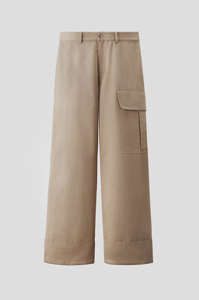 A pair of cargo trousers, presented FRONT, is crafted from a cotton weave, featuring a zip fly and button closure at the waist and integrated side seam pockets. A large patch leg pocket with a flap is positioned on the right leg, and the trousers are designed in a loose fit with wide, folded-up hems. - 1