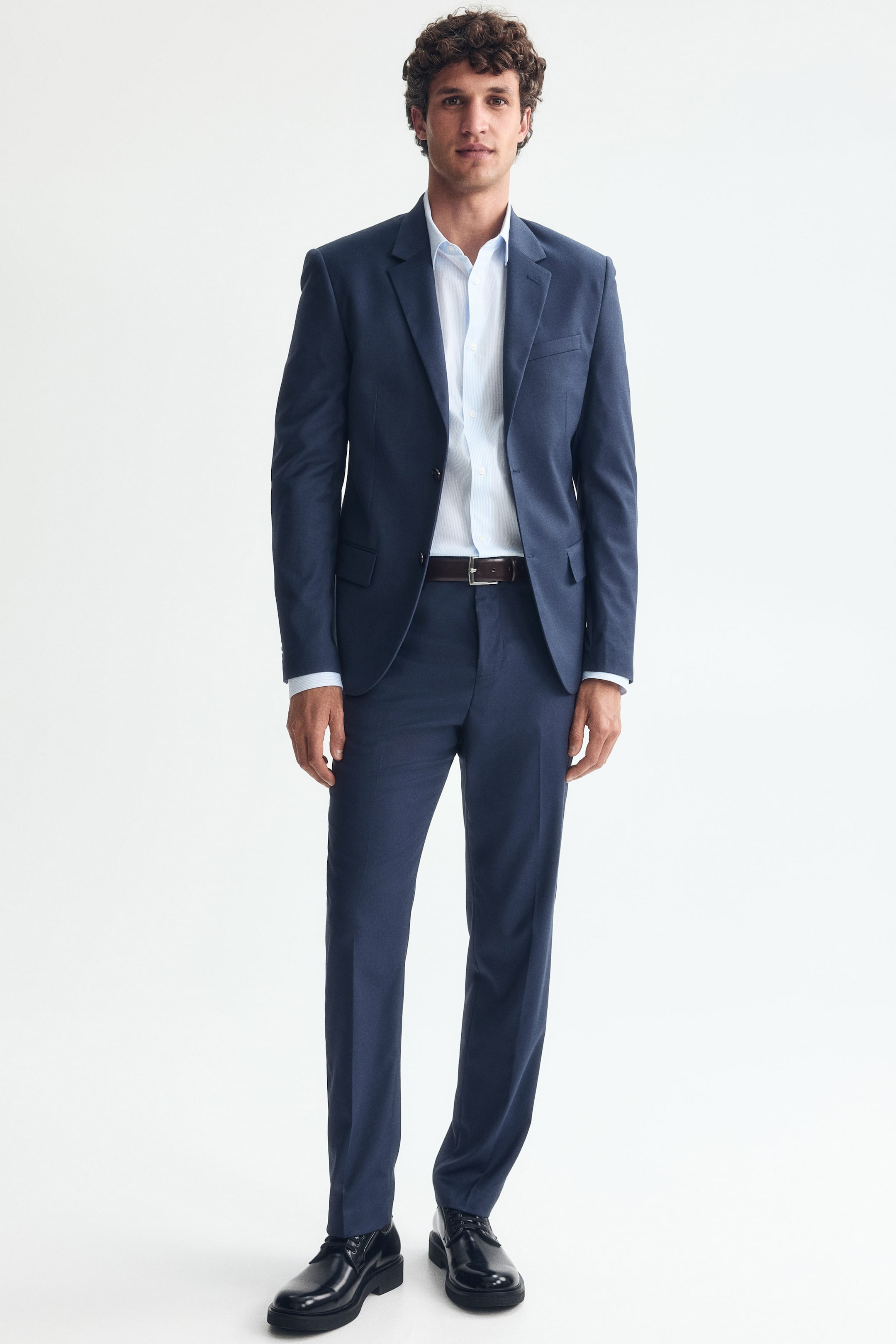 Ampliar la imagen: El modelo lleva puestos pantalones de traje azul oscuro, que presentan un corte ajustado y pliegues visibles a lo largo de las piernas. Los pantalones se combinan con un saco de traje a juego, una camisa azul claro, un cinturón oscuro y zapatos de vestir negros.