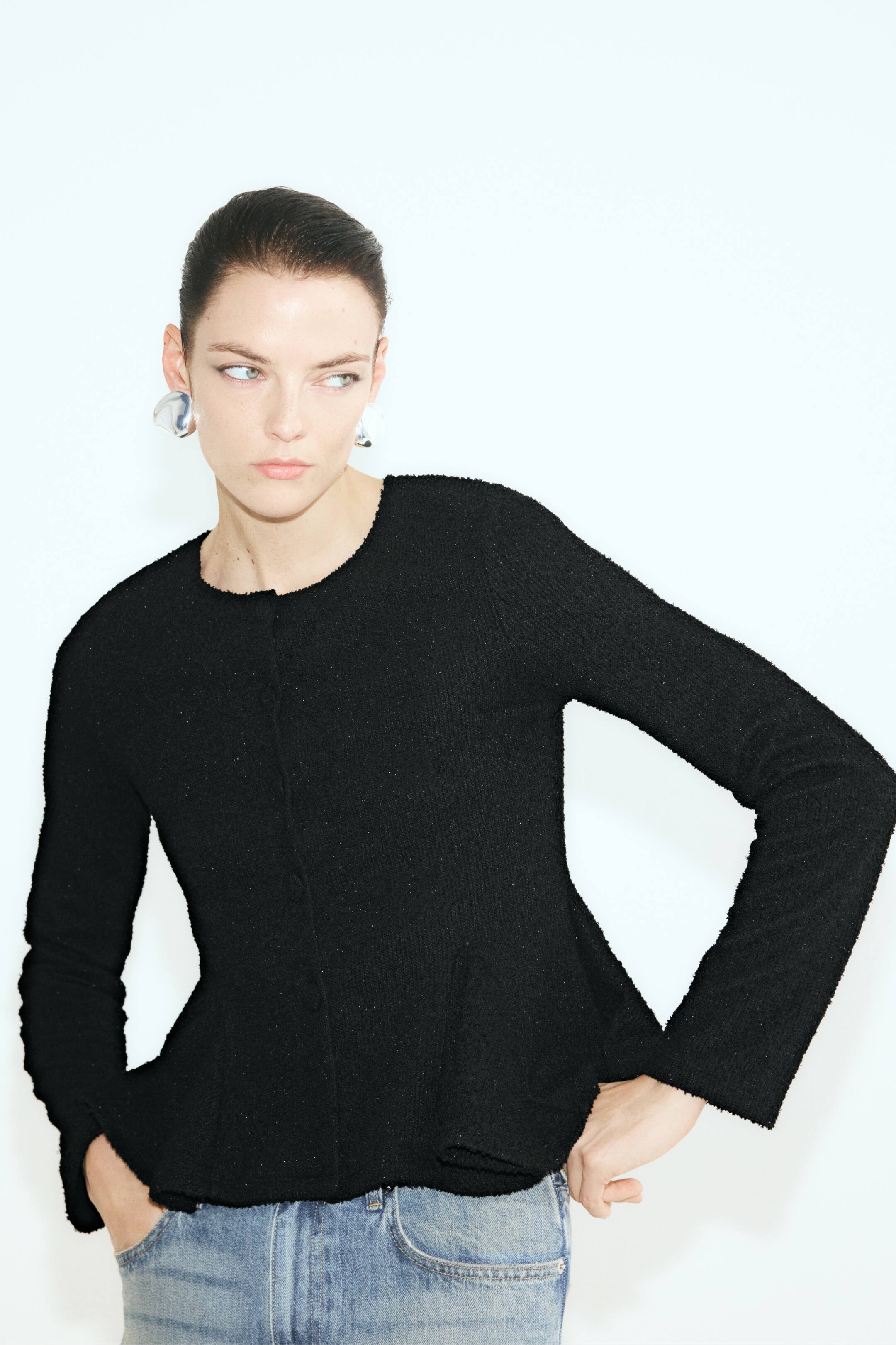 Visualizza l'immagine ingrandita: Una cardigan in jersey bouclé nera aderente, con scollo rotondo, maniche lunghe e un orlo peplum svasato con sottili fili glitterati, indossata dalla modella. La cardigan è abbinata a jeans in denim blu, con la modella che tiene le mani appoggiate vicino ai fianchi.