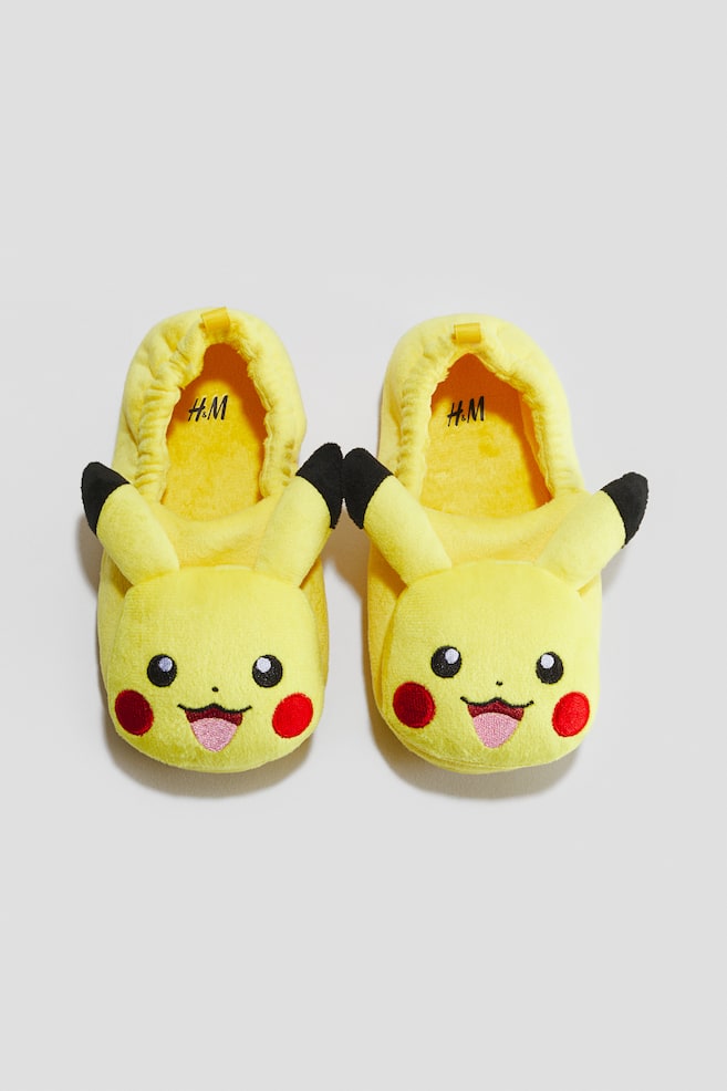 Soft slippers in a bright yellow plush fabric are resting side-by-side in a front-facing, top-down view. Each slipper is featuring a cartoon character face on the toe box with stuffed ears tipped in black, red circular cheeks, embroidered eyes, and an elasticated opening with a small pull tab at the heel. - 1