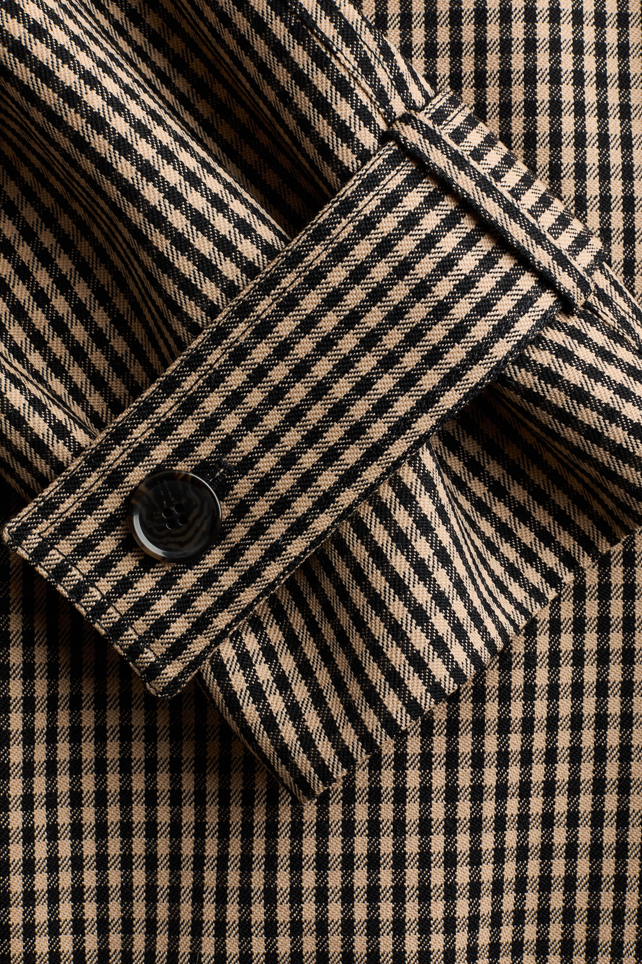 View larger image: A black and tan gingham check fabric featuring a strap detail with a dark mottled button. The fabric shows visible folds and creases, creating a textured appearance.