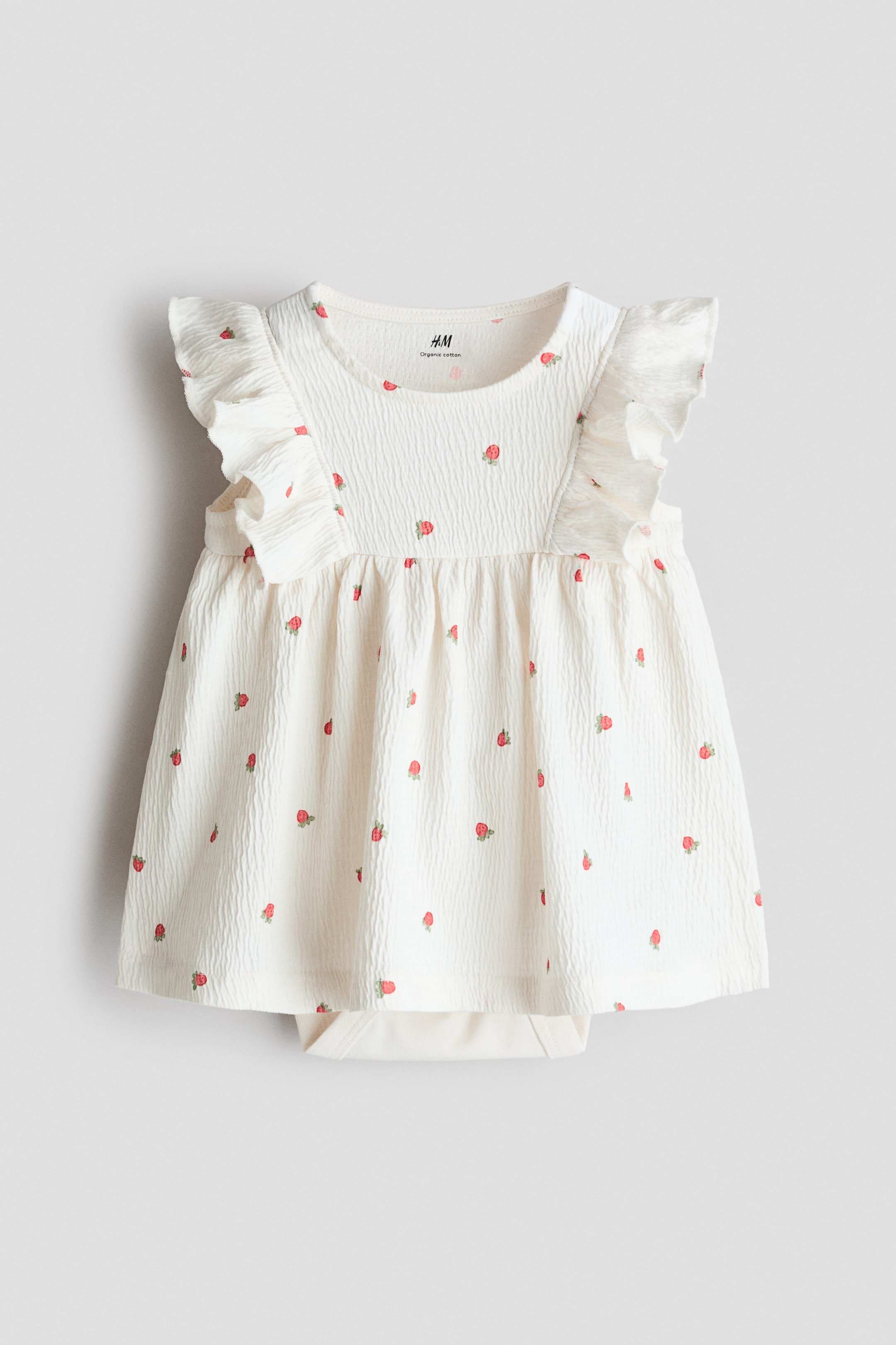 Agrandir l'image: Une robe pour bébé est présentée de face, montrant un tissu froissé blanc cassé orné de petites fraises rouges. Elle se distingue par un col rond, des manches courtes à volants et une jupe froncée, avec un body intégré blanc cassé visible sous l'ourlet.