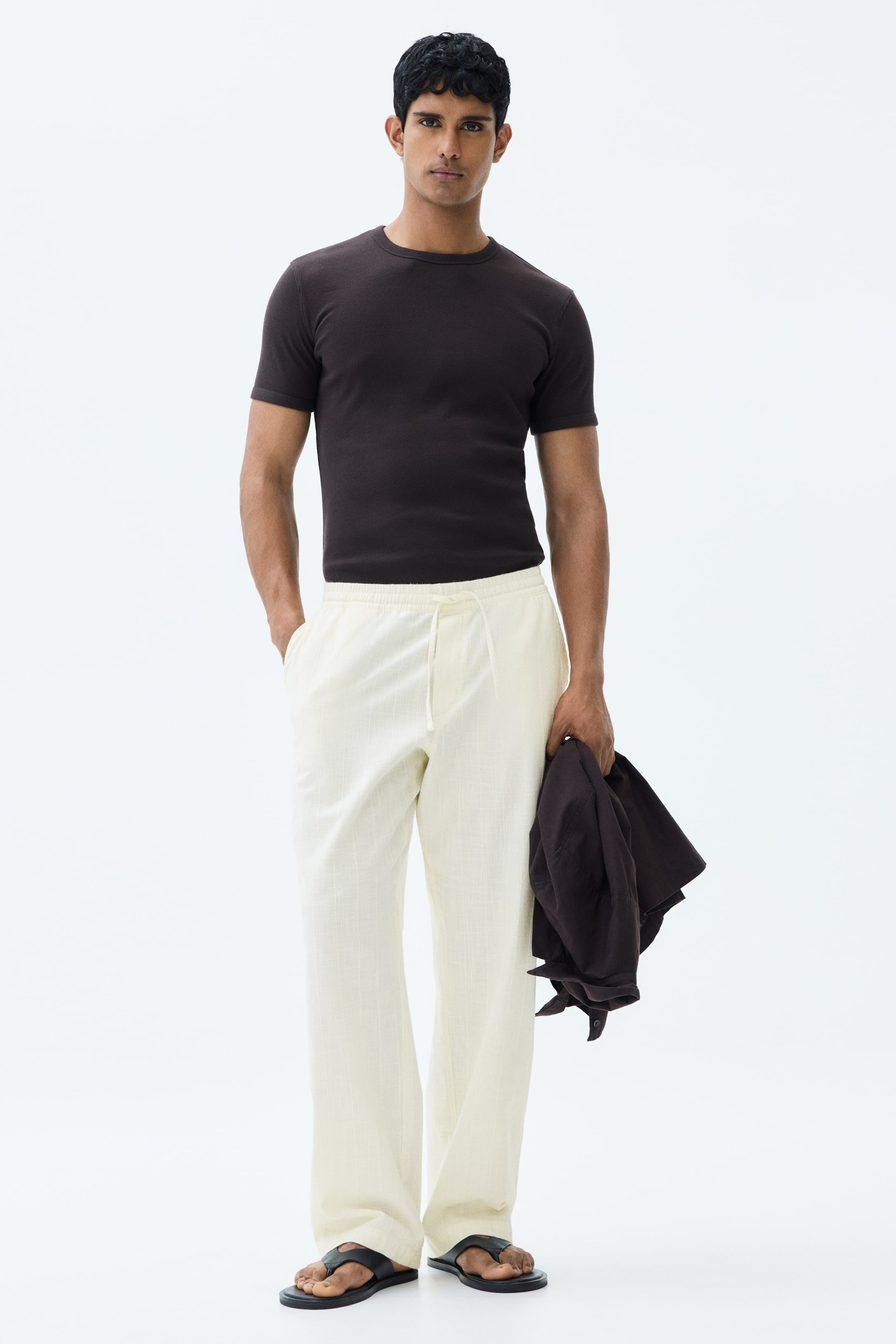 View larger image: The model is seen wearing trousers crafted from a light beige, airy cotton weave, offering a relaxed fit. These trousers come with a covered elasticated waist, complemented by a drawstring tie and diagonal side pockets. The model's right hand is casually placed in one of the pockets.