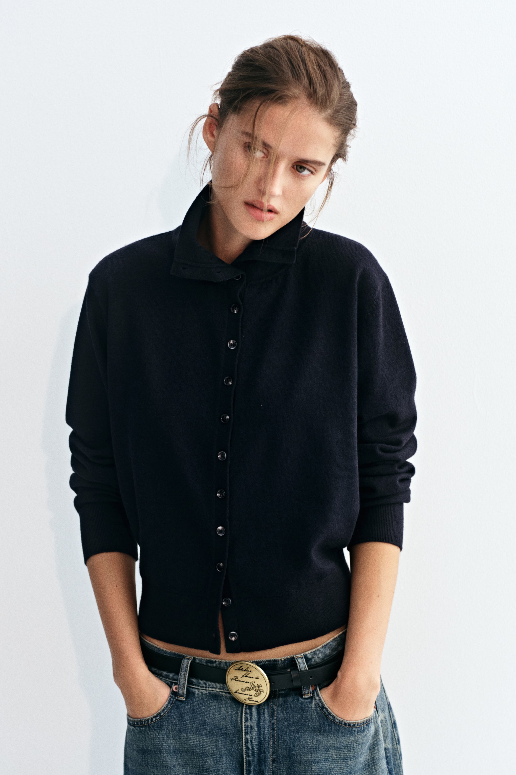 View larger image: A dark blue fine-knit cardigan featuring a high collar, button-down front, and ribbed cuffs is worn by the model. The cardigan is styled with blue jeans and a black belt with a round gold buckle.