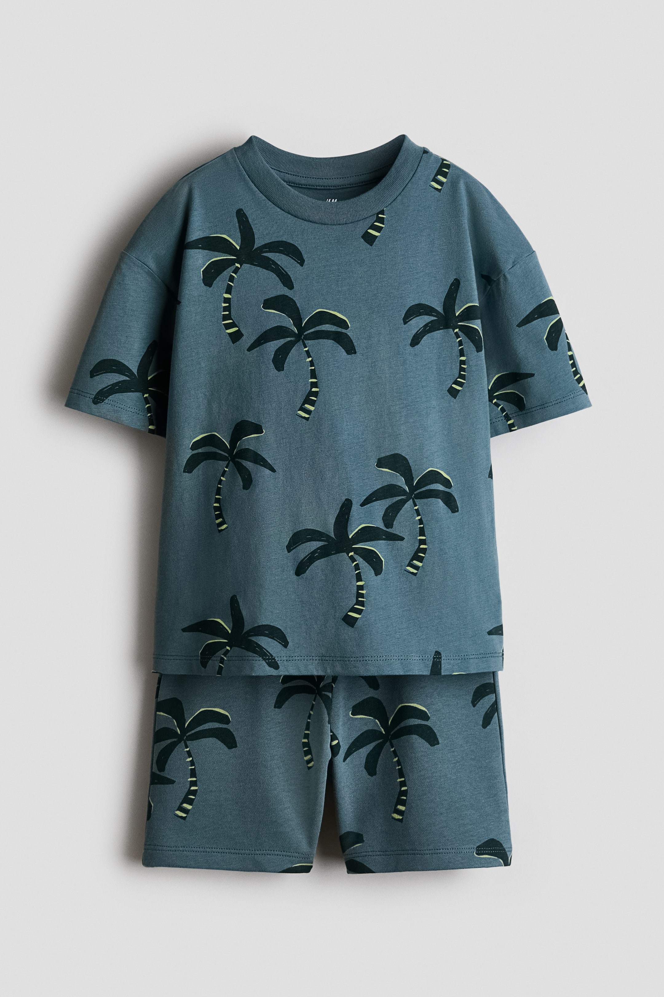 Agrandir l'image: Un ensemble de vêtements bleu-vert en deux pièces est présenté de face, comprenant un t-shirt à manches courtes et un short assorti, tous deux ornés de motifs de palmiers vert foncé. Le t-shirt en jersey de coton est conçu avec un col côtelé et des épaules tombantes, tandis que le short en tissu molleton léger affiche une taille élastiquée avec un cordon de serrage visible.