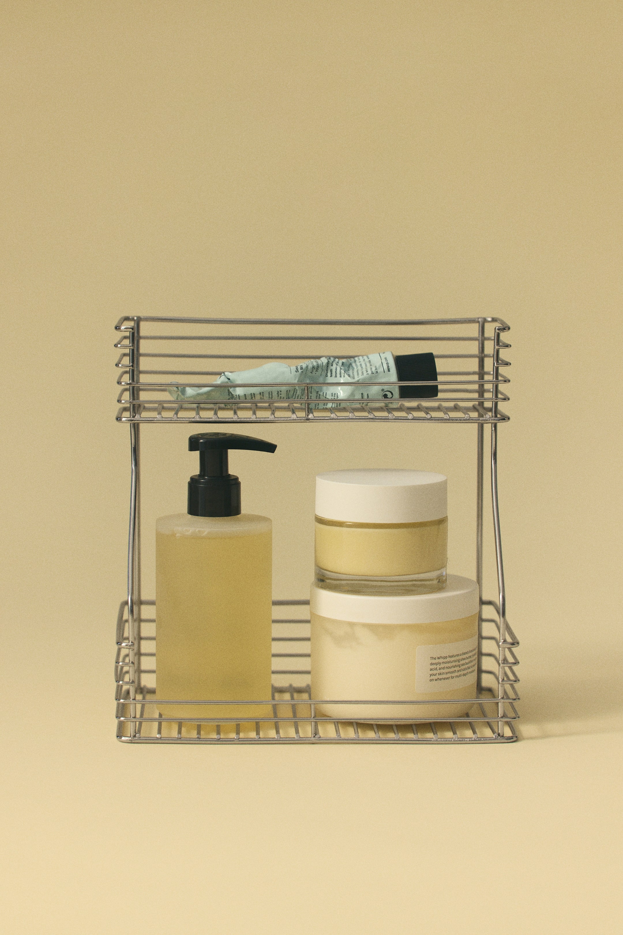 View larger image: A stainless steel wire shelving unit is standing upright, shown from a front-facing angle with a two-tier design featuring a wider bottom shelf and a narrower top shelf. The unit holds a yellow liquid pump bottle alongside two stacked cosmetic jars on the lower shelf, and a squeezed tube lying flat on the upper shelf.