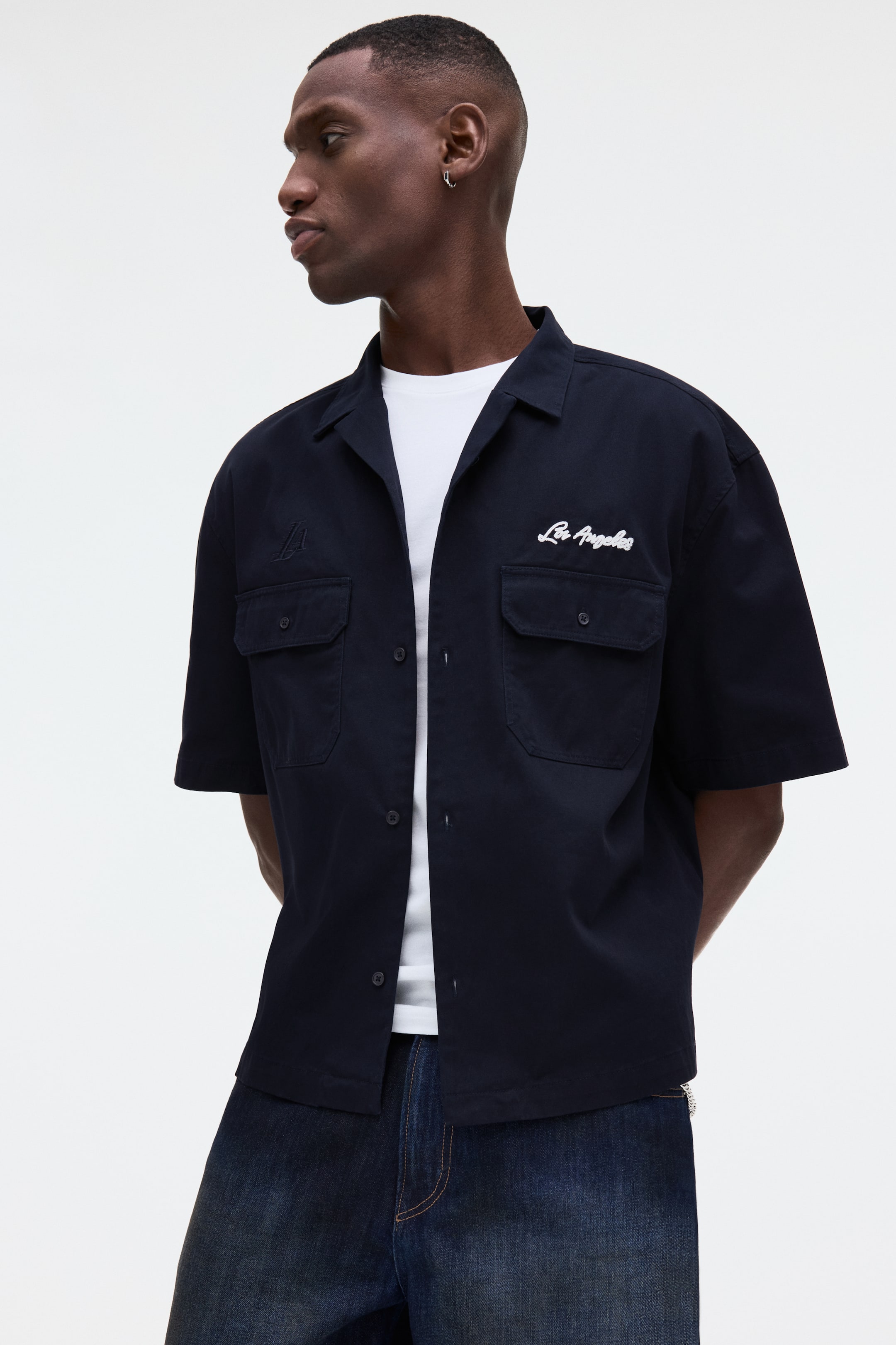 View larger image: A dark blue short-sleeved shirt is worn open over a white t-shirt, angled slightly to the right. It features a camp collar, a button-up front, and two chest flap pockets. The right chest displays white embroidered "Los Angeles" text above the pocket, while a stylized "A" logo appears on the left chest above the pocket.