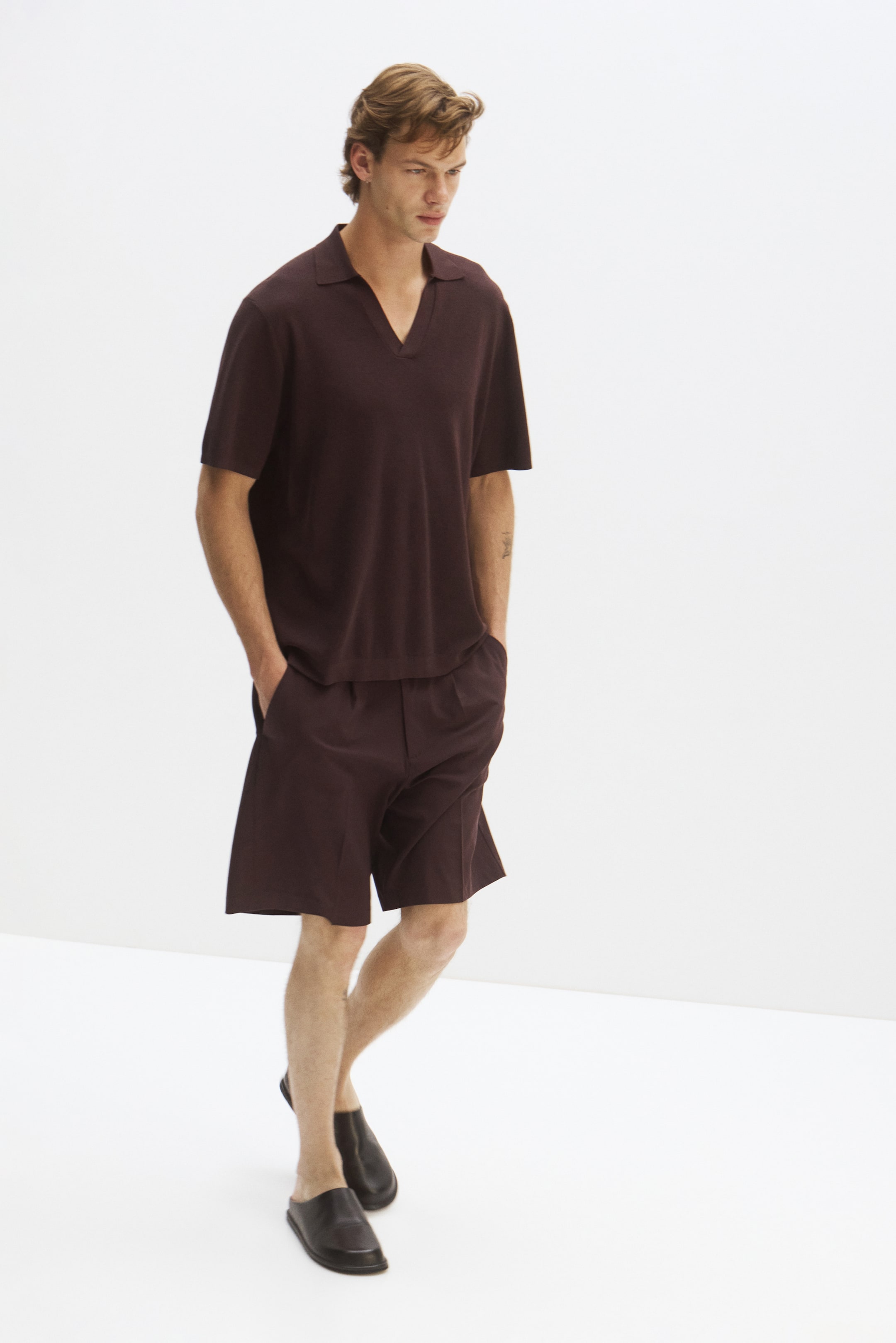 Grotere afbeelding bekijken: Een donkerbruine gebreide poloshirt en bijpassende op maat gemaakte shorts worden gepresenteerd op een model dat naar voren en iets naar rechts kijkt. Het poloshirt heeft een gespreide kraag en een open v-hals sluiting, terwijl de relaxed-fit shorts zijzakken hebben en boven de knie komen.