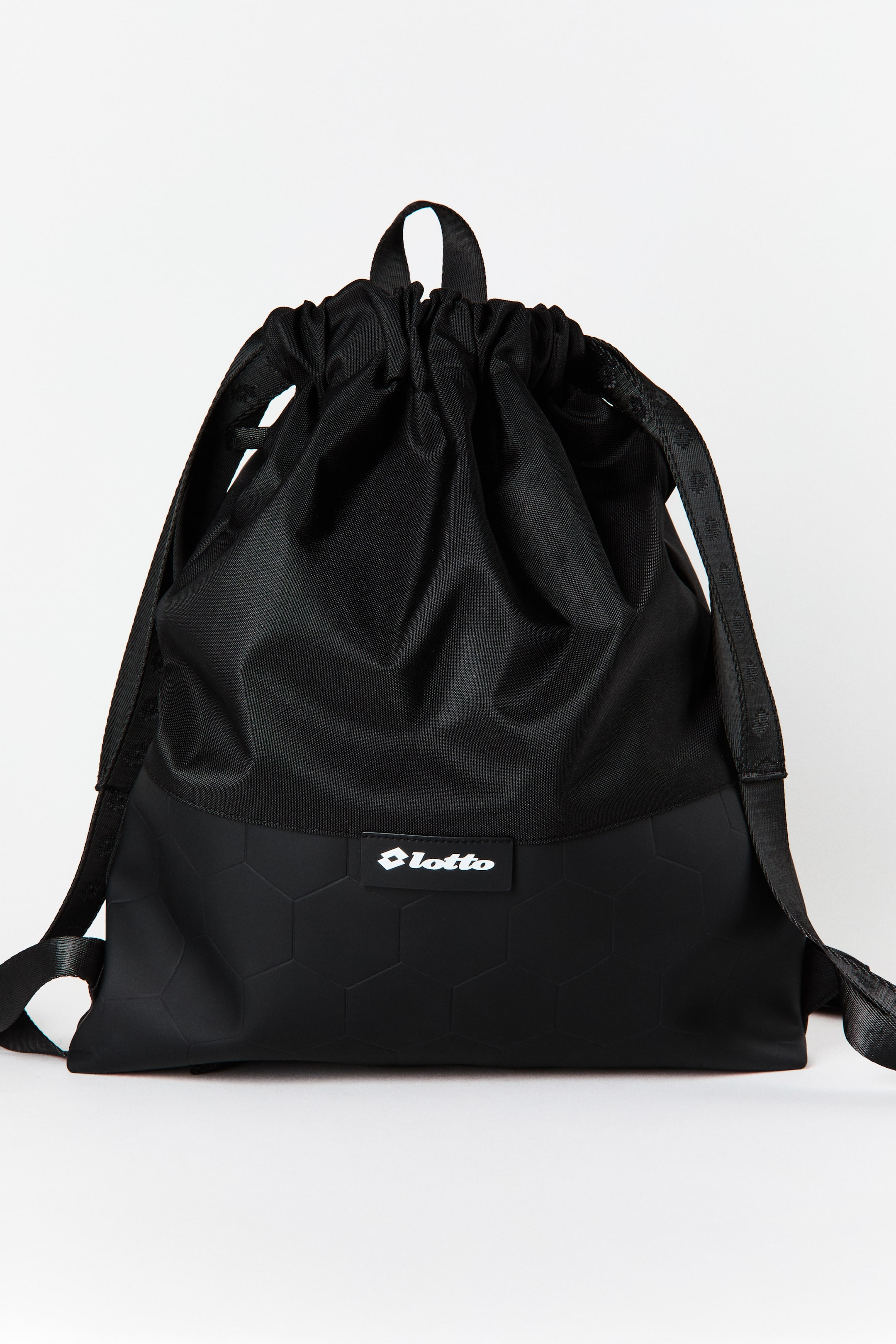 Ampliar la imagen: Una mochila negra con cordón está orientada hacia adelante, con una sección superior de tela recogida y una sección inferior con un patrón hexagonal en relieve. Un logotipo blanco de Lotto es visible en el frente, y las correas de hombro negras con un patrón sutil se extienden por los lados.