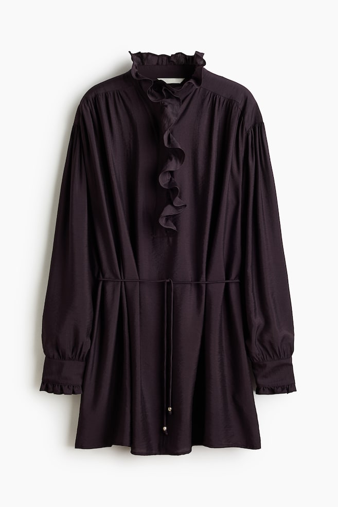 Ein dunkelviolettes Tunika-Kleid präsentiert seine Vorderseite, mit einem gerafften Stehkragen und einer vertikalen Rüsche, die über das Oberteil fließt. Es zeigt lange, voluminöse Ärmel mit gesmokten, gerafften Manschetten und einem Taillenband aus dem gleichen Stoff mit goldfarbenen Perlendetails. - 1