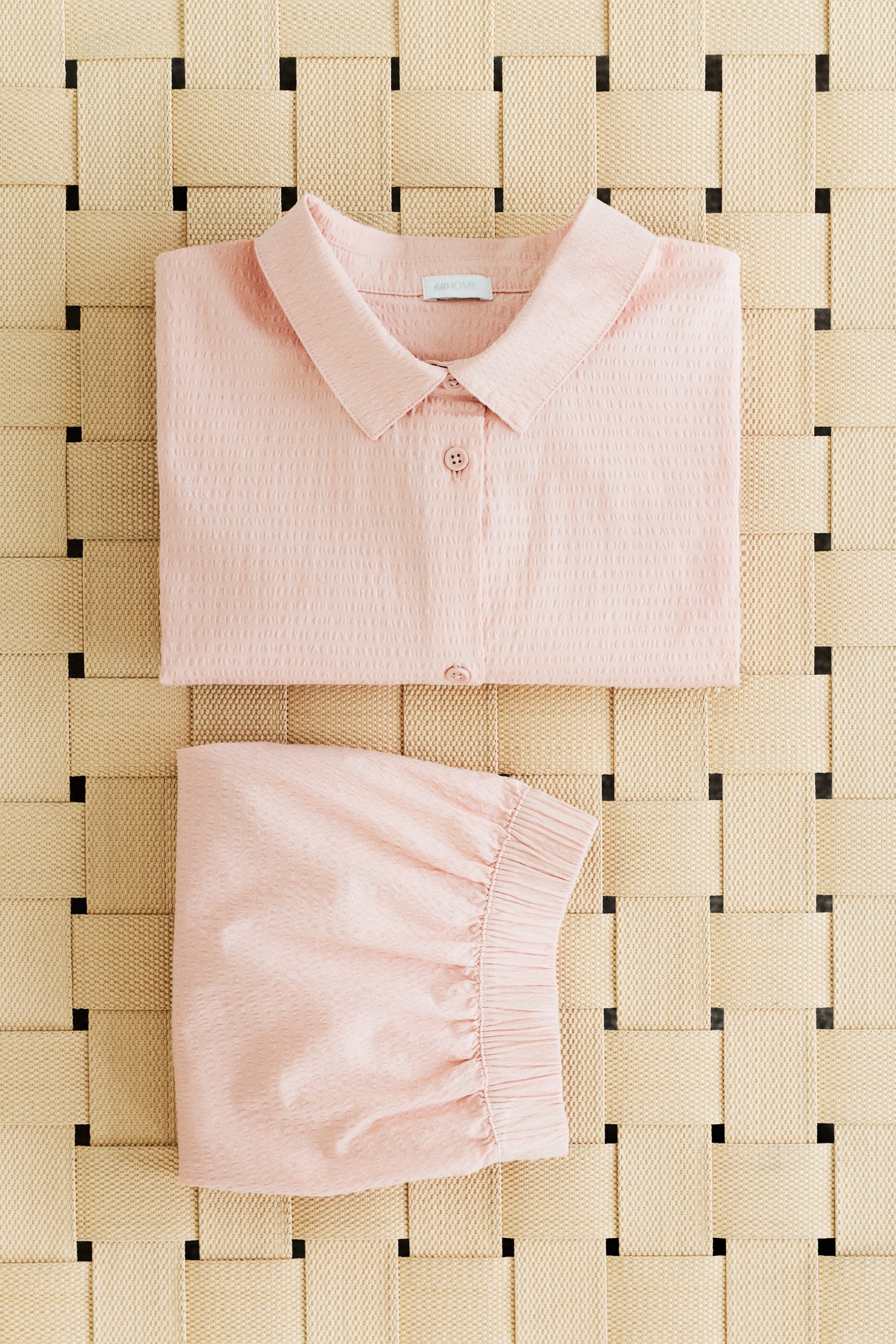 Visa större bild: Det rosa pyjamassetet, bestående av en skjorta och shorts i seersucker-tyg, visas med båda plaggen vikta. Skjortan visar sin FRAMSIDA med en nedvikt krage och synliga knappar framtill, medan shortsen visar sin FRAMSIDA med dold resår i midjan.