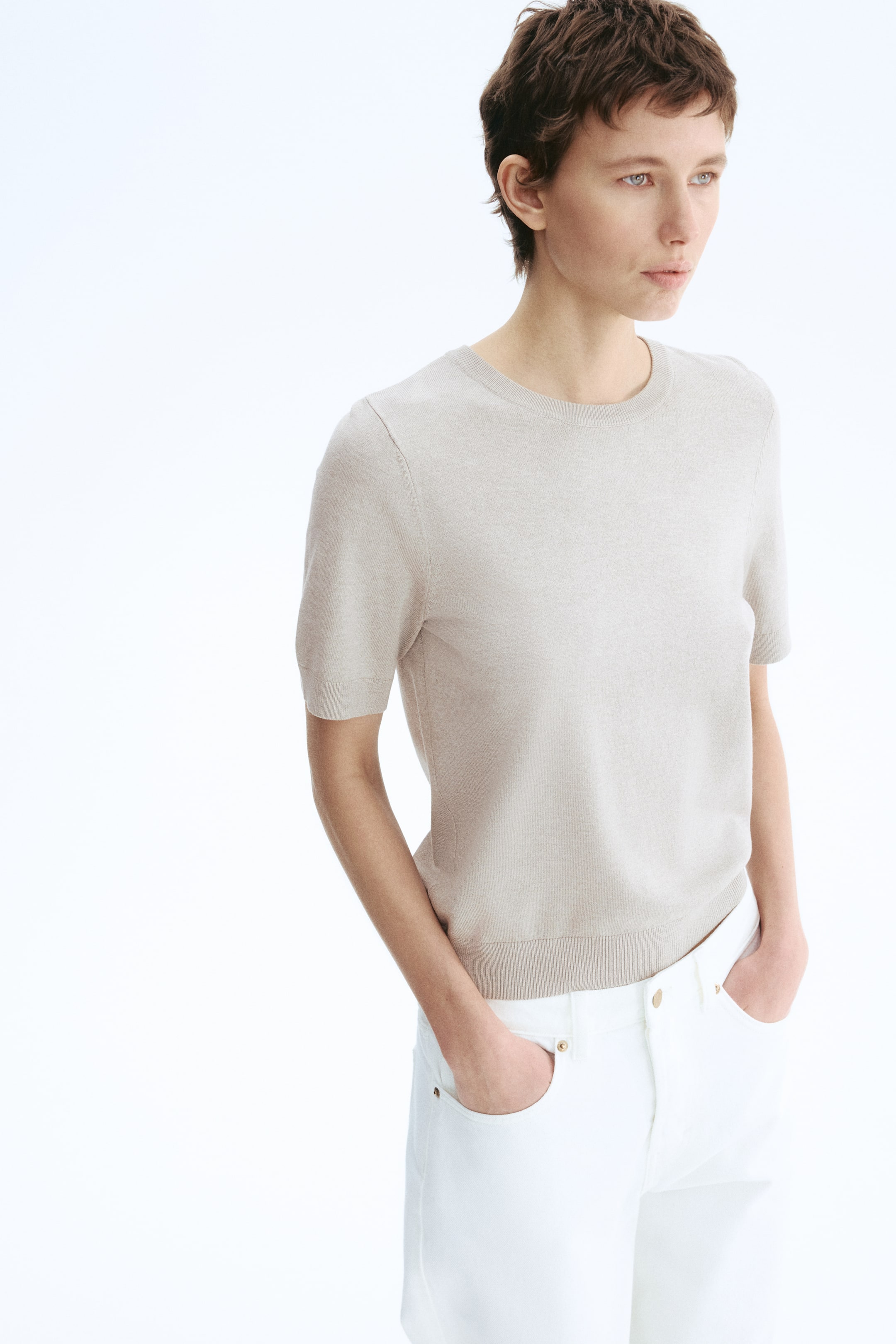 View larger image: A light beige fine-knit short-sleeved top is being worn, angled slightly to the right. It is featuring a crew neck, short sleeves with ribbed cuffs, and a ribbed bottom hem, fitting closely to the body.