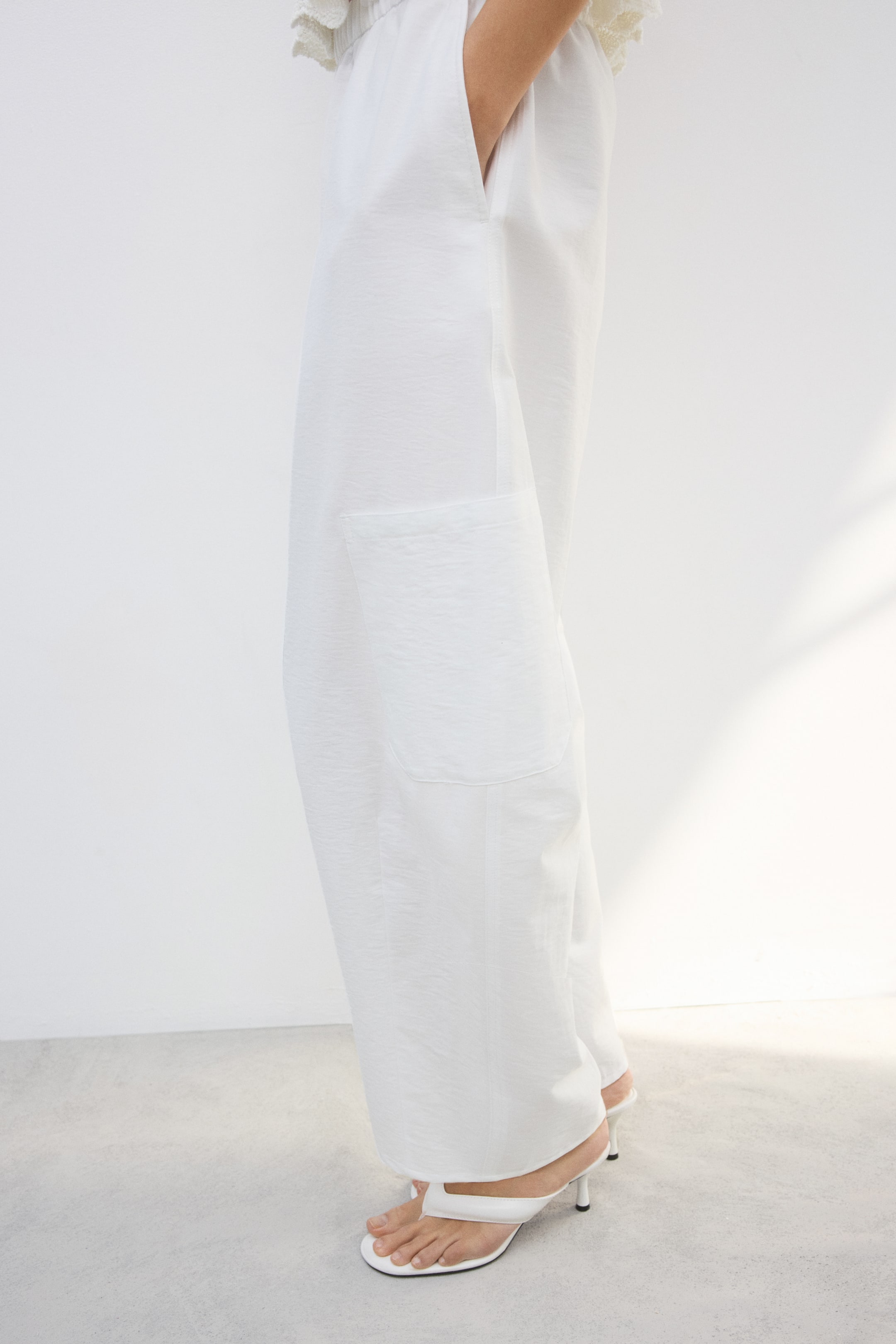 View larger image: White wide-leg pants are displayed from a side angle, showcasing a subtly textured fabric and a gathered waistband. A large patch pocket is visible on the left leg, while a hand rests in an open side pocket on the right leg.