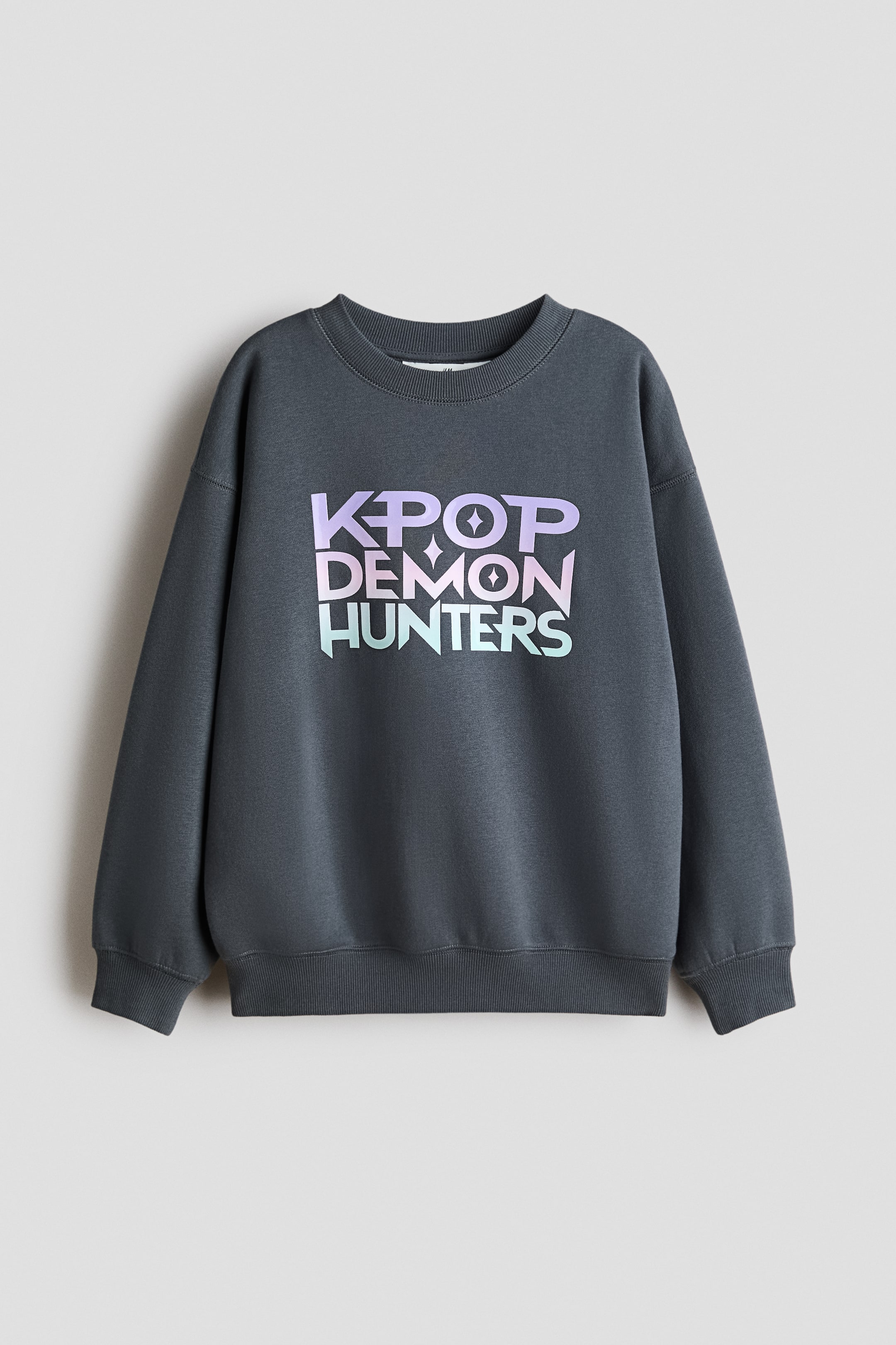 Ver imagen más grande: Vista frontal de una sudadera oversize en gris oscuro. Presenta un cuello redondo con acabado de canalé, hombros caídos y puños y bajo acanalados. En la parte delantera muestra el texto «K-POP DEMON HUNTERS» en un estampado degradado de morado a verde azulado.