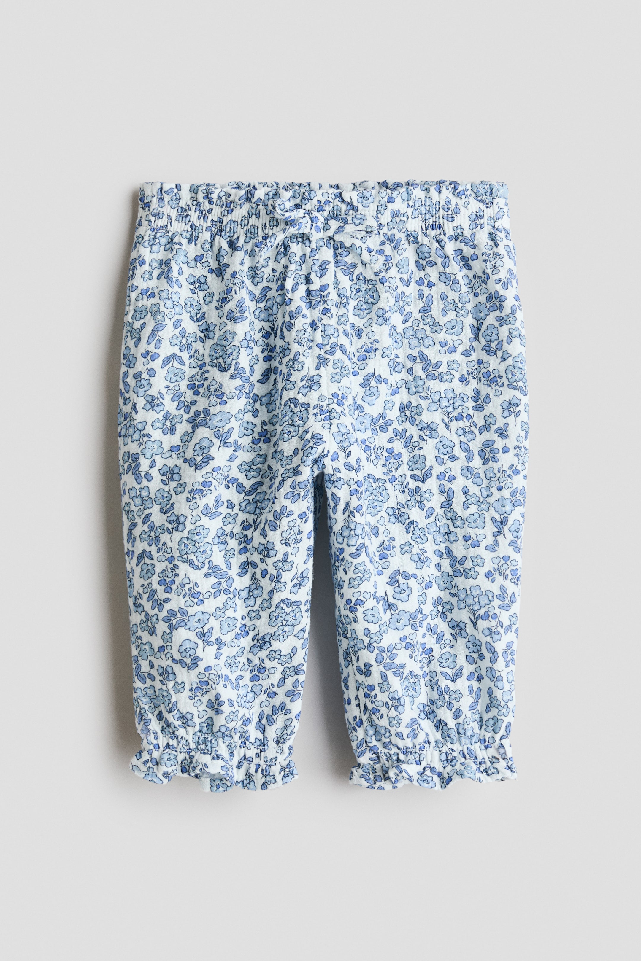 Prikaži veću sliku: Pantalone imaju belu podlogu s plavim cvetnim dezenom preko cele površine, elastičan pojas s malom mašnom sprijeda i nabrane, volanaste manžete na gležnjevima.