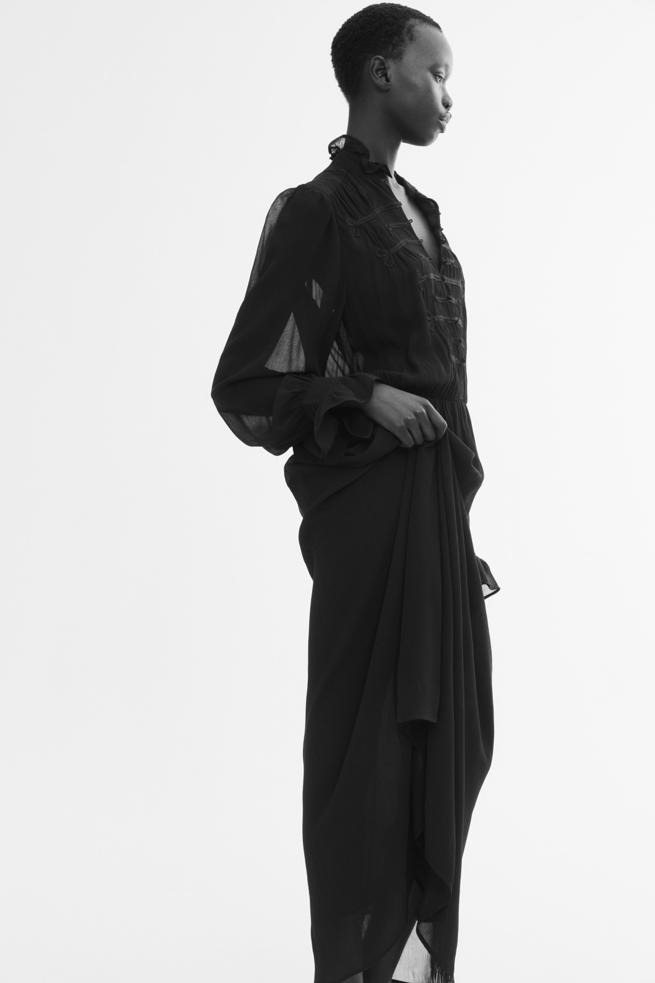 Größeres Bild ansehen: Ein schwarzes, halbtransparentes Kleid mit Rüschenkragen, strukturierter Oberpartie und langem, drapiertem Rock wird vom Model getragen. Das Model hält seitlich einen Teil des fließenden Rockstoffs.