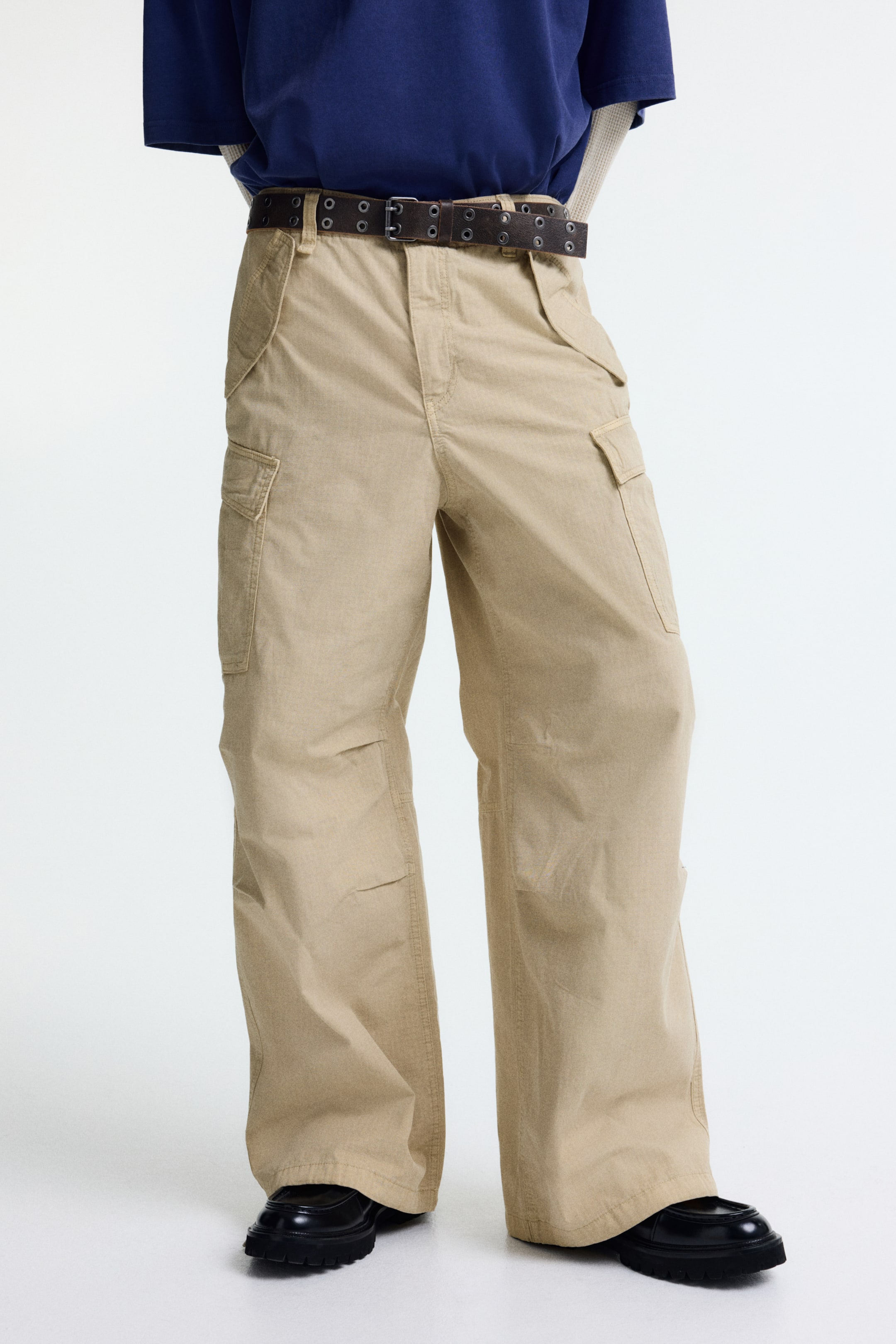 拡大画像を表示: カーキのワイドレッグカーゴパンツは、フロントポケットと脚の外側に配置された大きなフラップカーゴポケットが付いており、ルーズフィットを提供しています。ダークブラウンのダブルグロメットベルトは、ベルトループを通して着用され、ウエストを締めています。