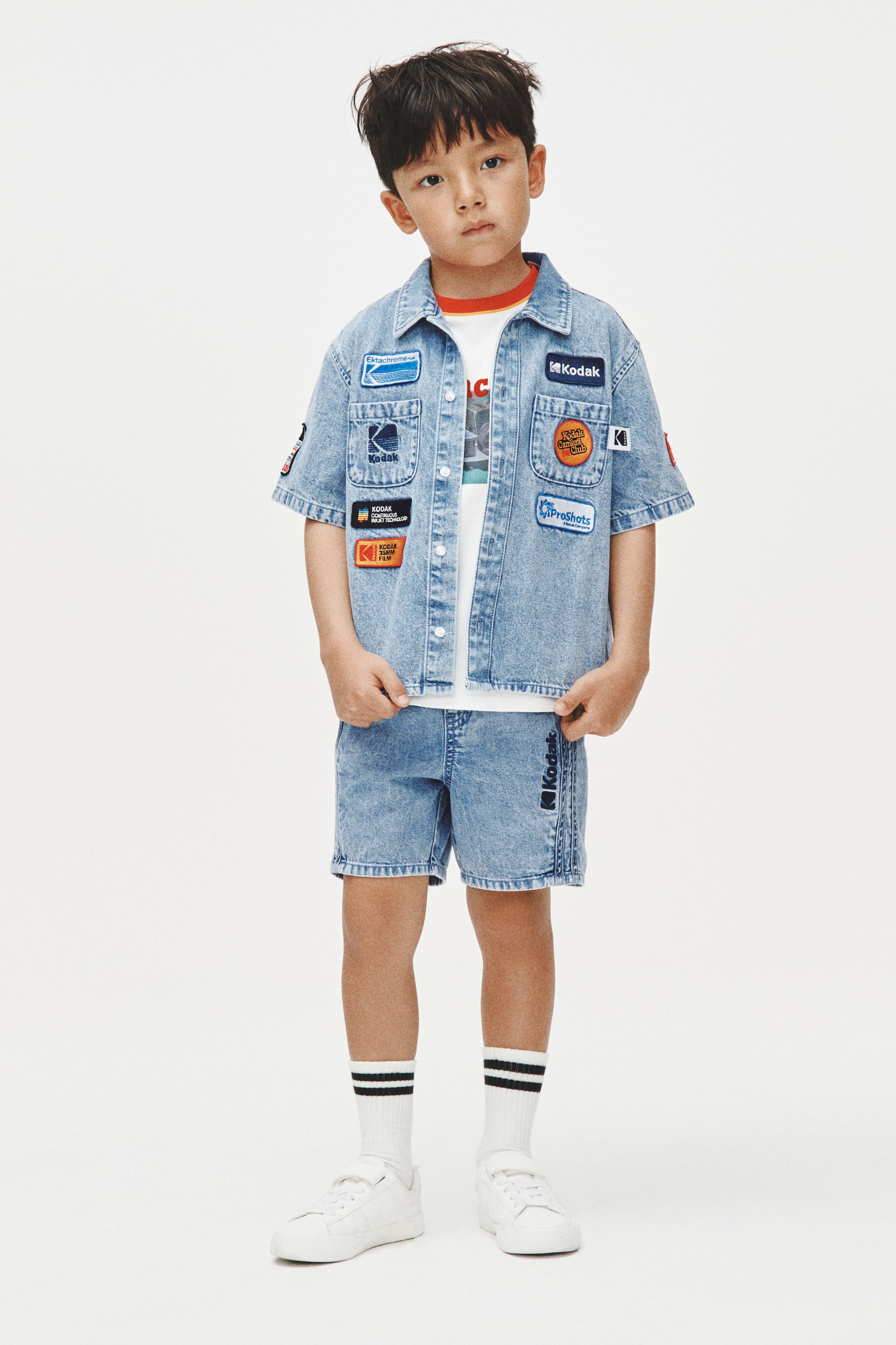 View larger image: "A light blue denim short-sleeved shirt na may mga makukulay na brand patches, dalawang chest pockets, at isang classic collar, ay isinusuot nang bukas sa ibabaw ng isang puting t-shirt. Ang kaparehong light blue denim shorts ay may brand logo na naka-print nang patayo sa gilid."