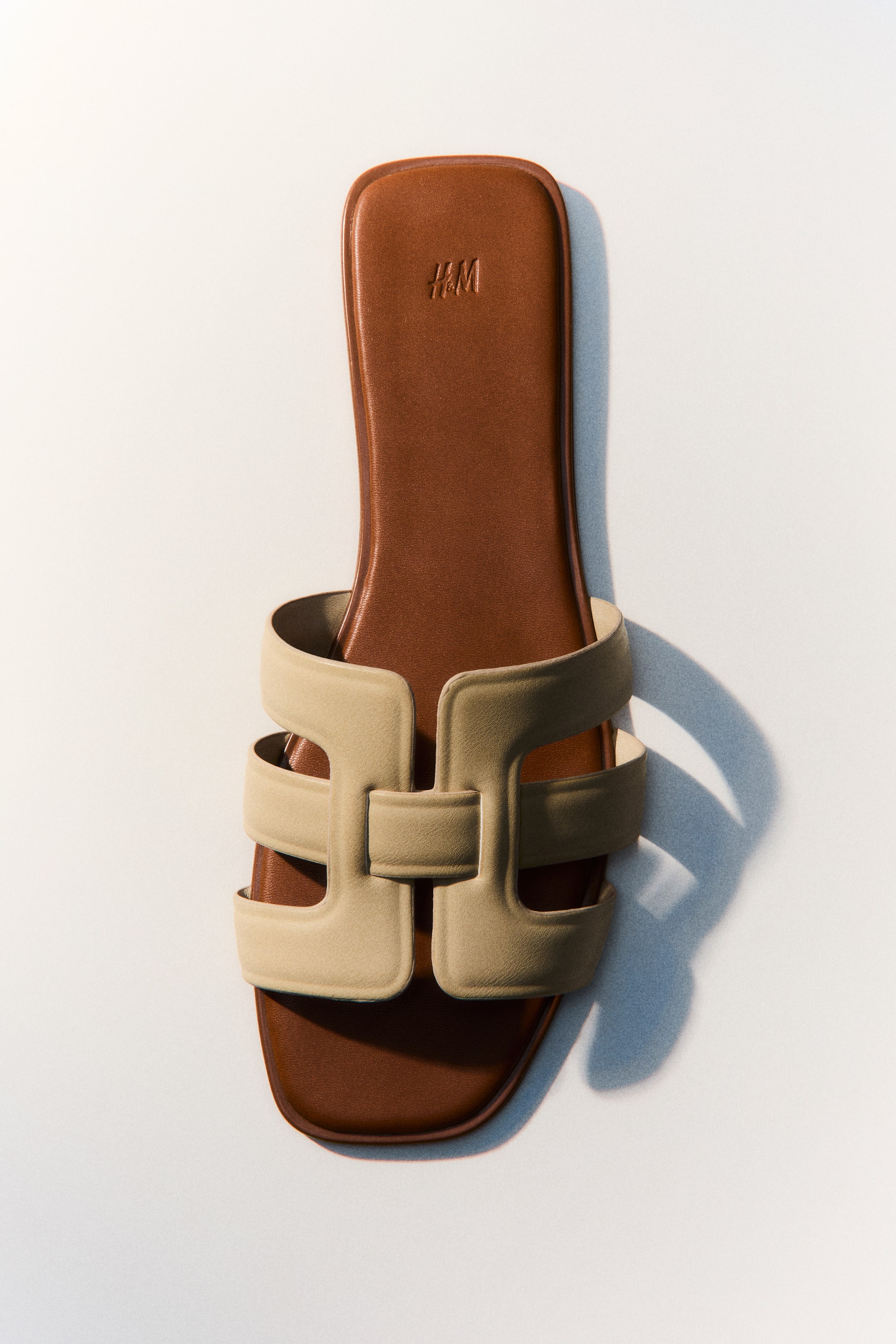 Größeres Bild ansehen: Ein Slide-Sandal präsentiert ein braunes Fußbett mit einem H&M-Logo, das oben eingeprägt ist. Ein breiter, beigefarbener H-förmiger Riemen bildet das Obermaterial.