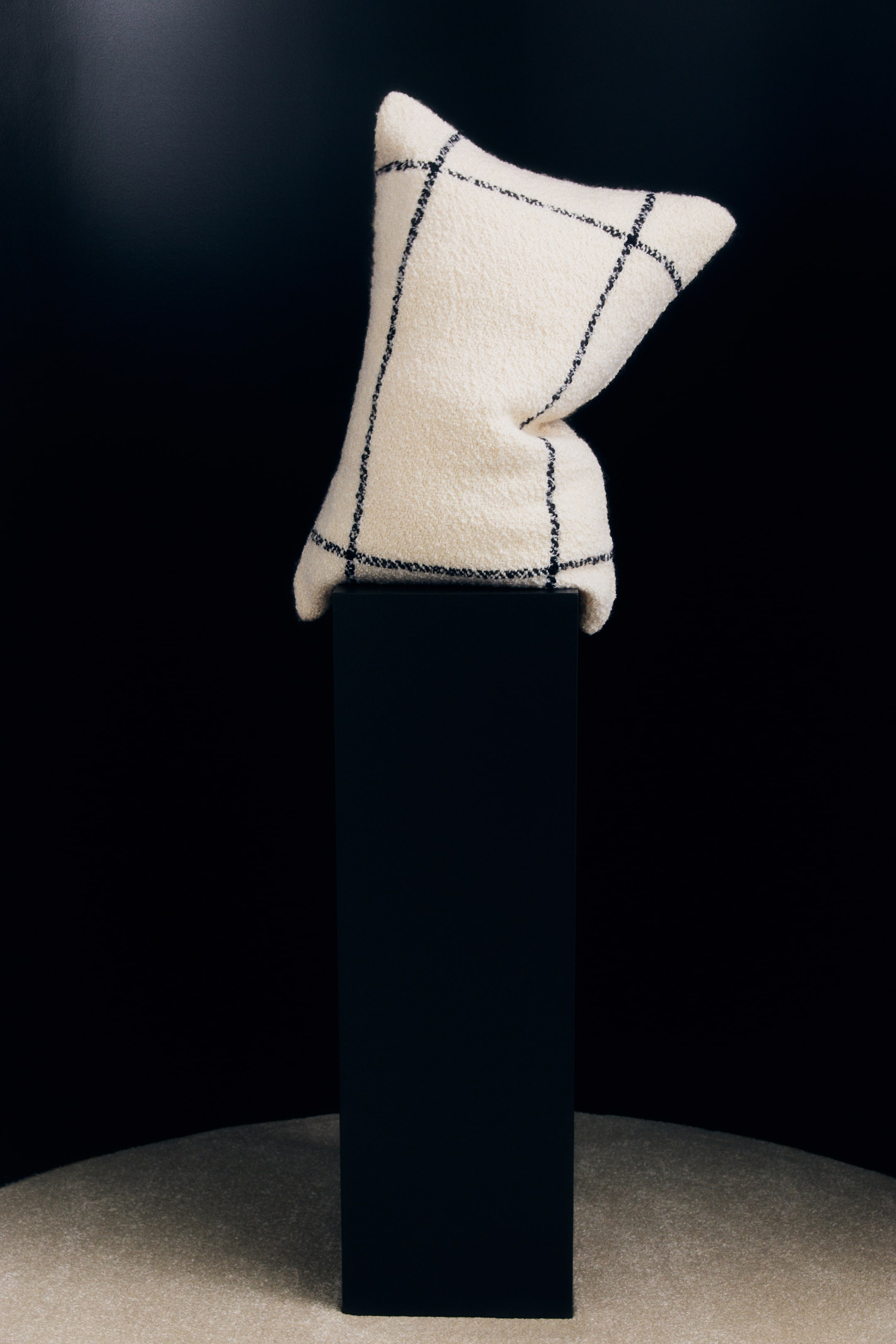 Vezi imaginea mai mare: O pernă decorativă off-white, cu textură bouclé, cu model în carouri tip windowpane negru, este sprijinită vertical pe o coloană neagră, evidențiind designul frontal și forma sa moale, ușor pliată.