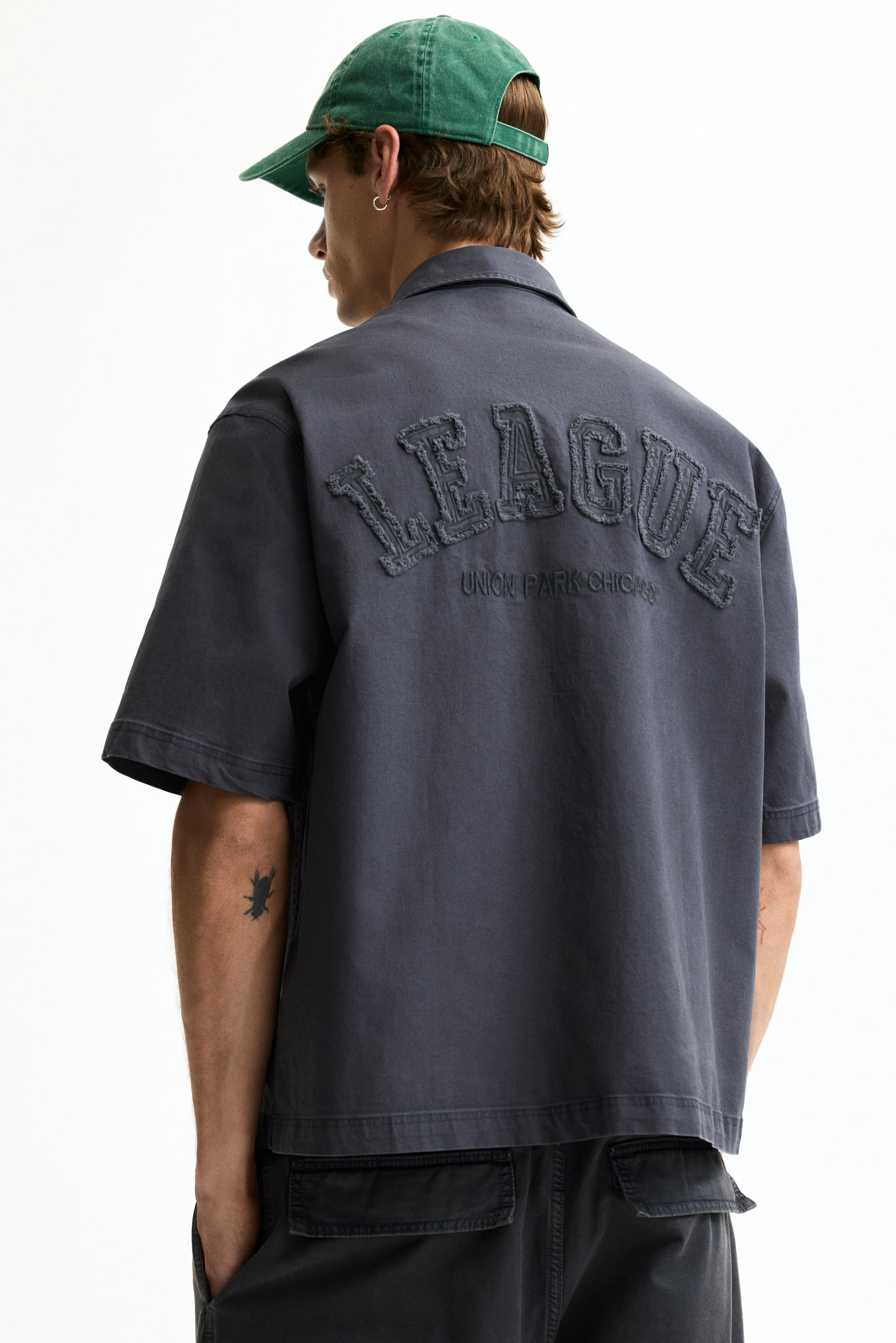 Ampliar la imagen: Una camisa gris oscuro de manga corta con cuello está de espaldas, vista desde atrás, mostrando "LEAGUE" en grandes letras aplicadas y desgastadas, y "UNION PARK CHICAGO" en texto bordado más pequeño debajo.